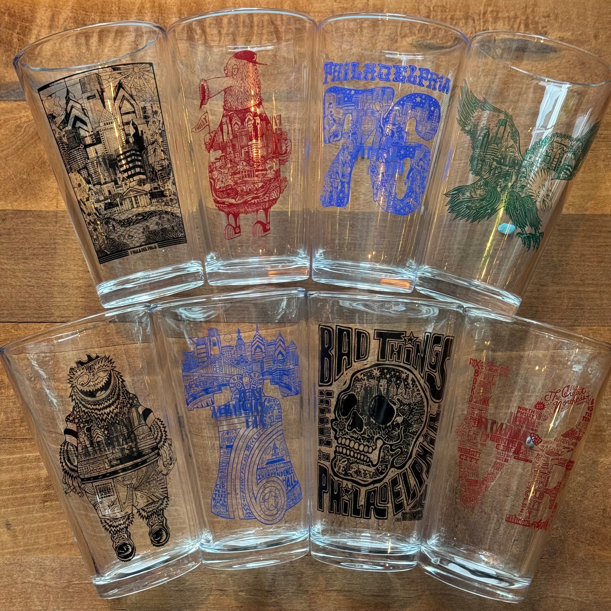 A collection of eight Paul Carpenter Philly Pint Glasses each with a different design related to Philadelphia's landmarks and skyline, displayed on a wooden surface.