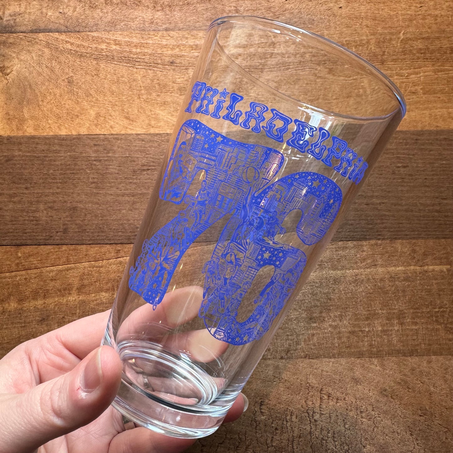 A hand holding an empty pint glass with blue Philadelphia-themed skyline decorative patterns - Philly Pint Glasses from Paul Carpenter.