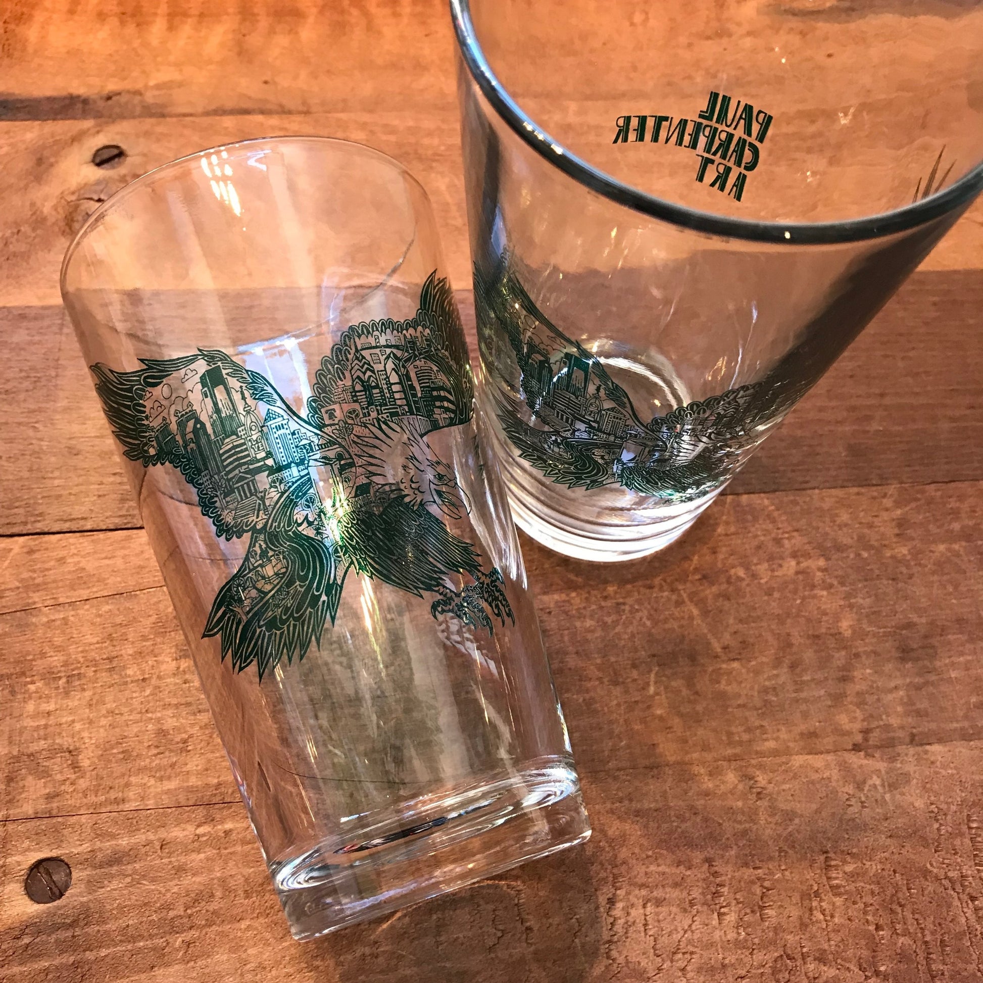 Two empty Paul Carpenter Philly Pint Glasses with eagle designs, one standing upright and the other tilted, on a wooden surface.