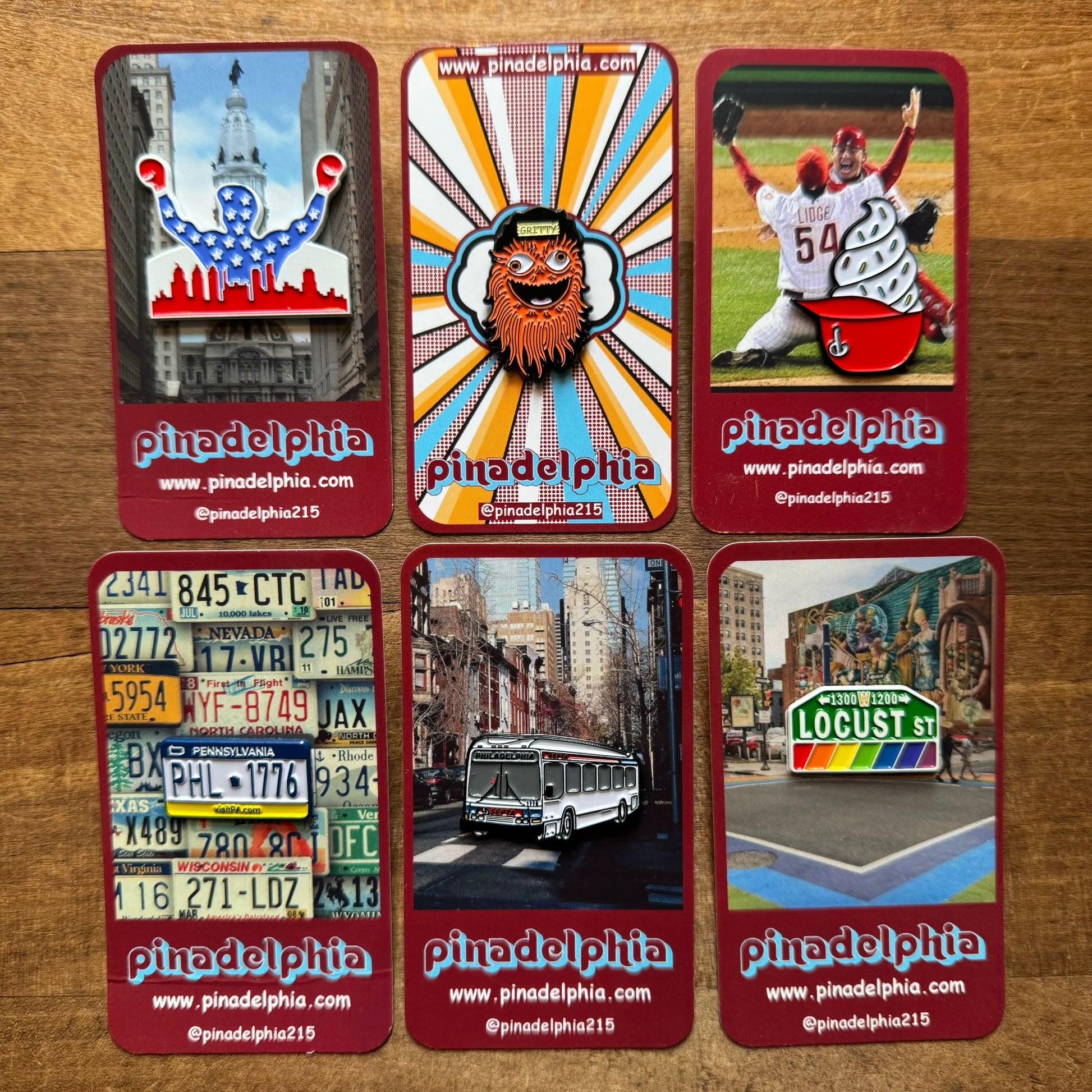 Six Pinadelphia Philly-Themed Enamel Pins II featuring symbols and scenes from Philly pride displayed on a wooden surface.