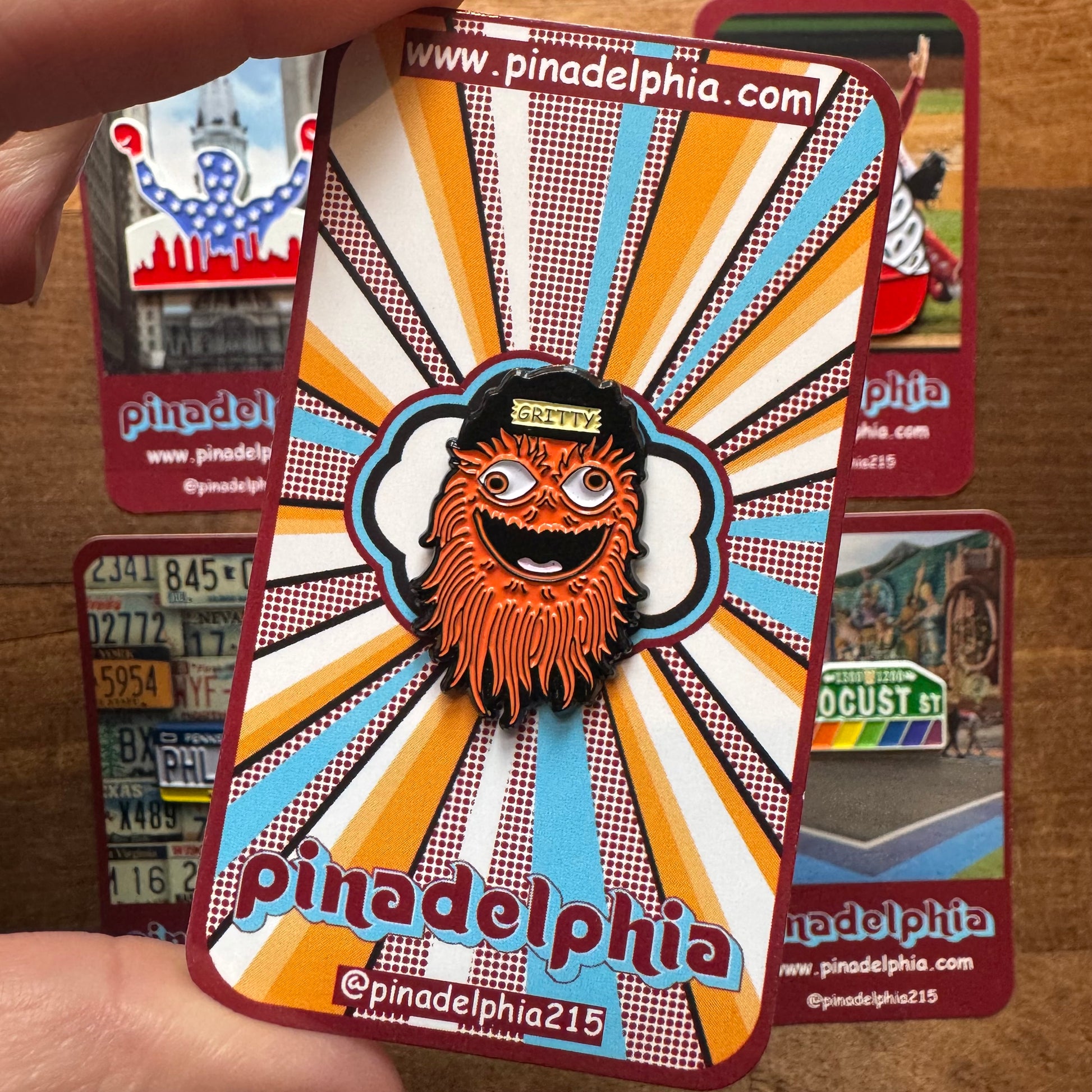 A hand holding a Pinadelphia Philly-Themed Enamel Pin II celebrating Philly pride, featuring a stylized beard and sunglasses design with "Pinadelphia" and a radial pattern in the background.