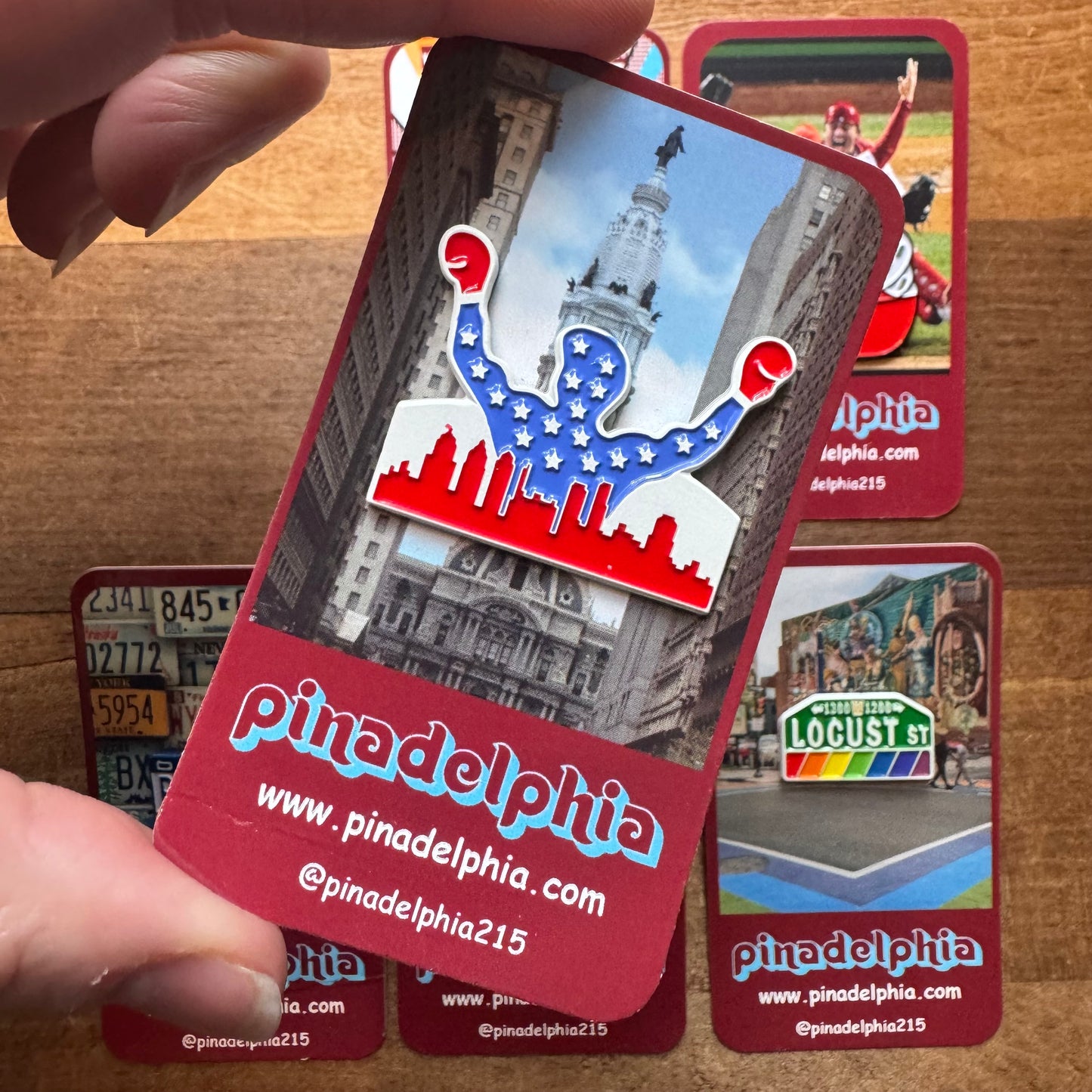 A hand holding an enamel pin-shaped business card with a Philly-Themed Enamel Pins II from Pinadelphia, showcasing the city's landmarks and the social media handles of the brand "Pinadelphia.