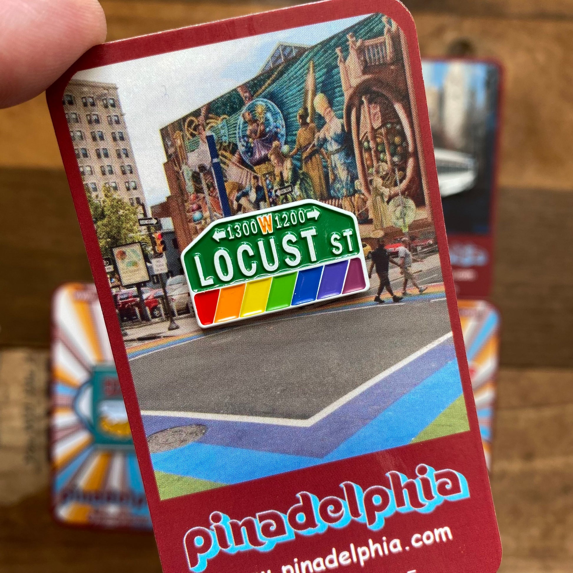 A person holding a colorful postcard featuring the street sign of locust st in Philadelphia with a vibrant city background, adorned with Pinadelphia's Philly-Themed Enamel Pins II.