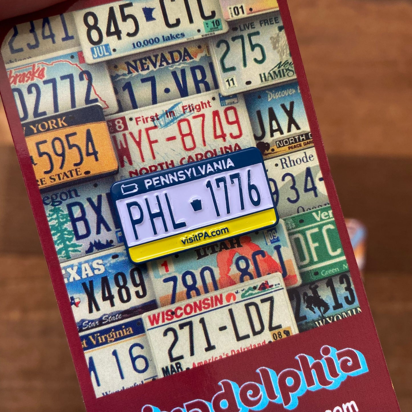 A hand holding a brochure with a collage of U.S. license plates and a Pennsylvania license plate shaped magnet, adorned with Philly pride enamel pins from Pinadelphia's Philly-Themed Enamel Pins II.