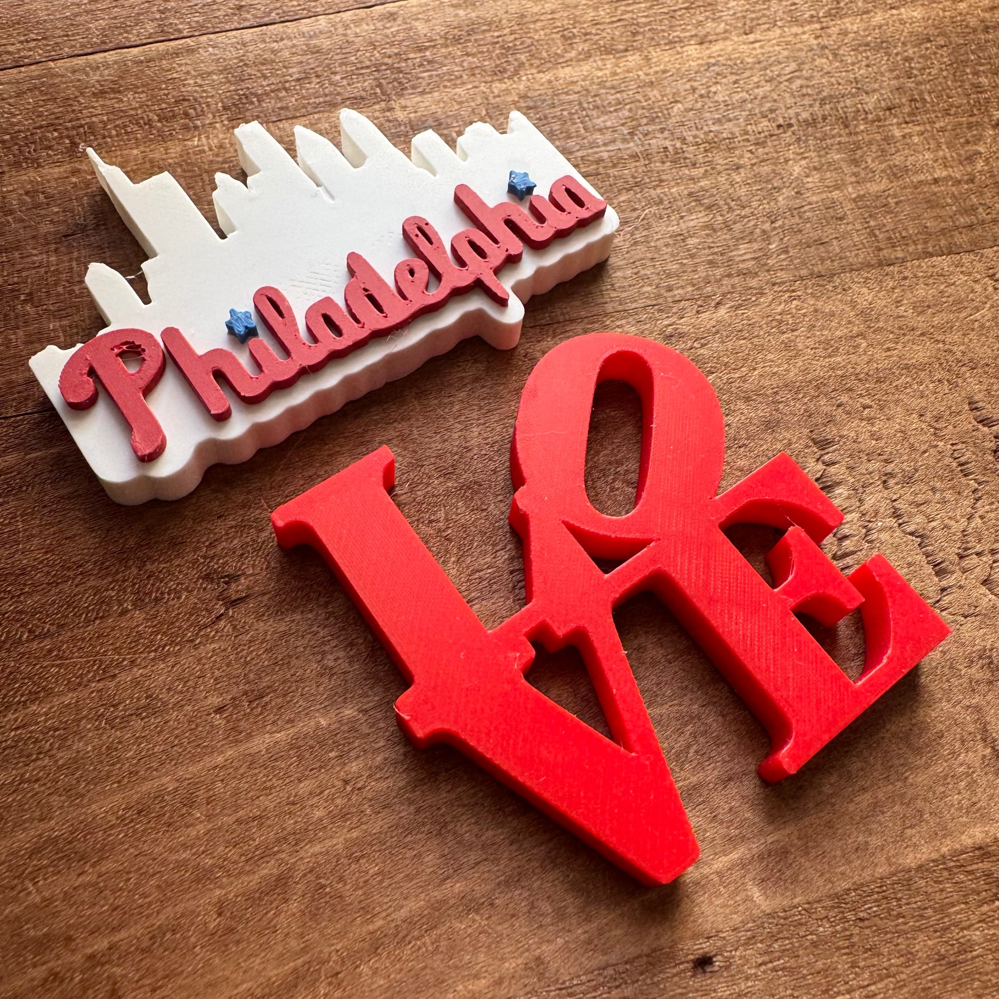 Two Philly Magnets on a wooden surface: one in the shape of the Philadelphia skyline, and the other spelling "love" in bold, red letters by Rosewood Home.