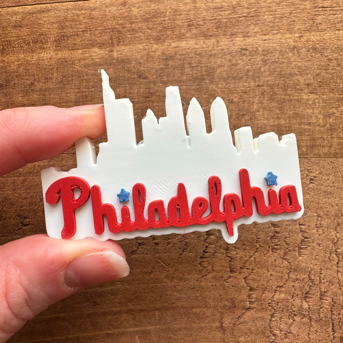 A hand holding a white and red Philly Magnet shaped like the Philadelphia skyline against a wooden background.