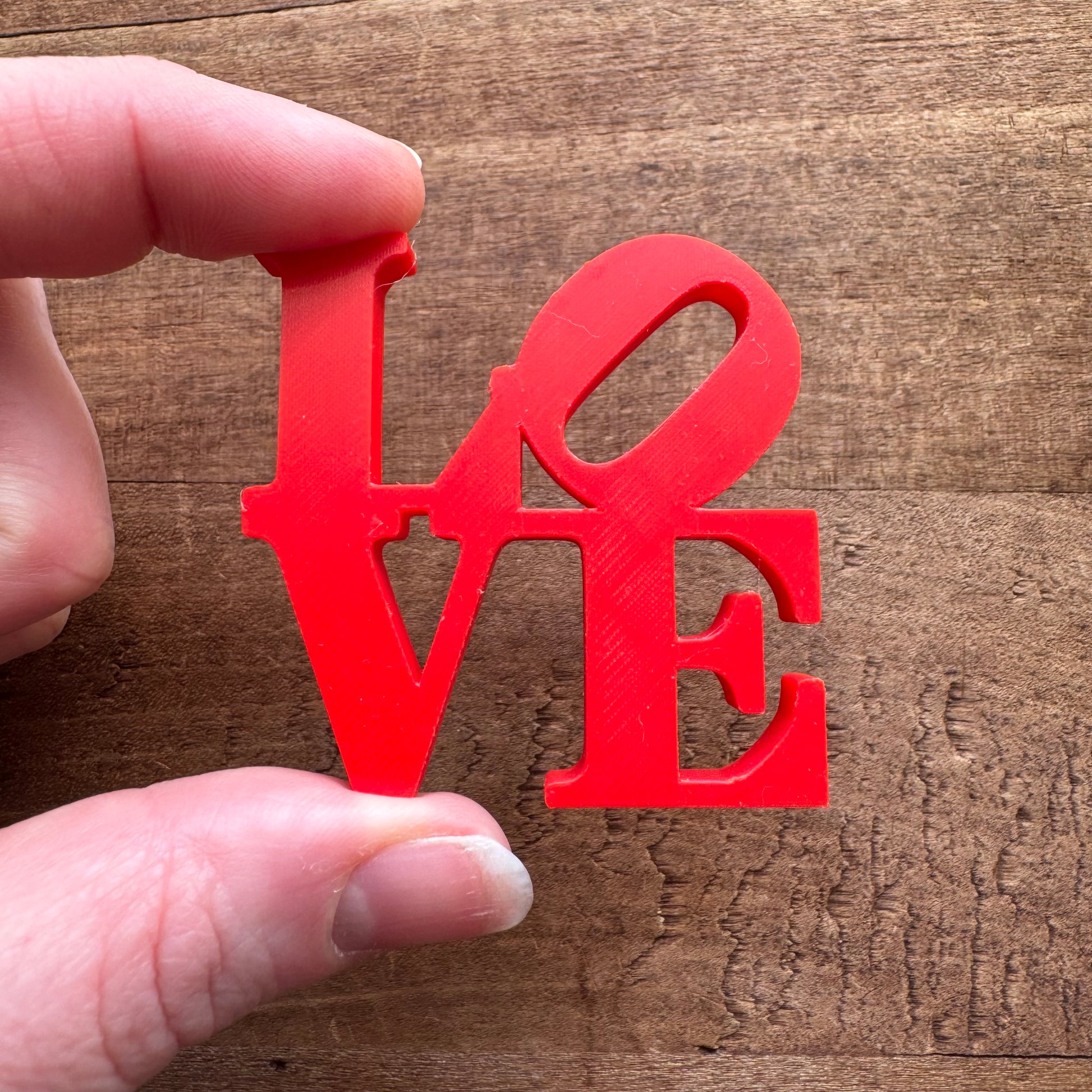 A hand holding a red, plastic cutout of the word "love," where the "o" is replaced with a Philly themed peace symbol, against a wooden background featuring Rosewood Home Philly Magnets.