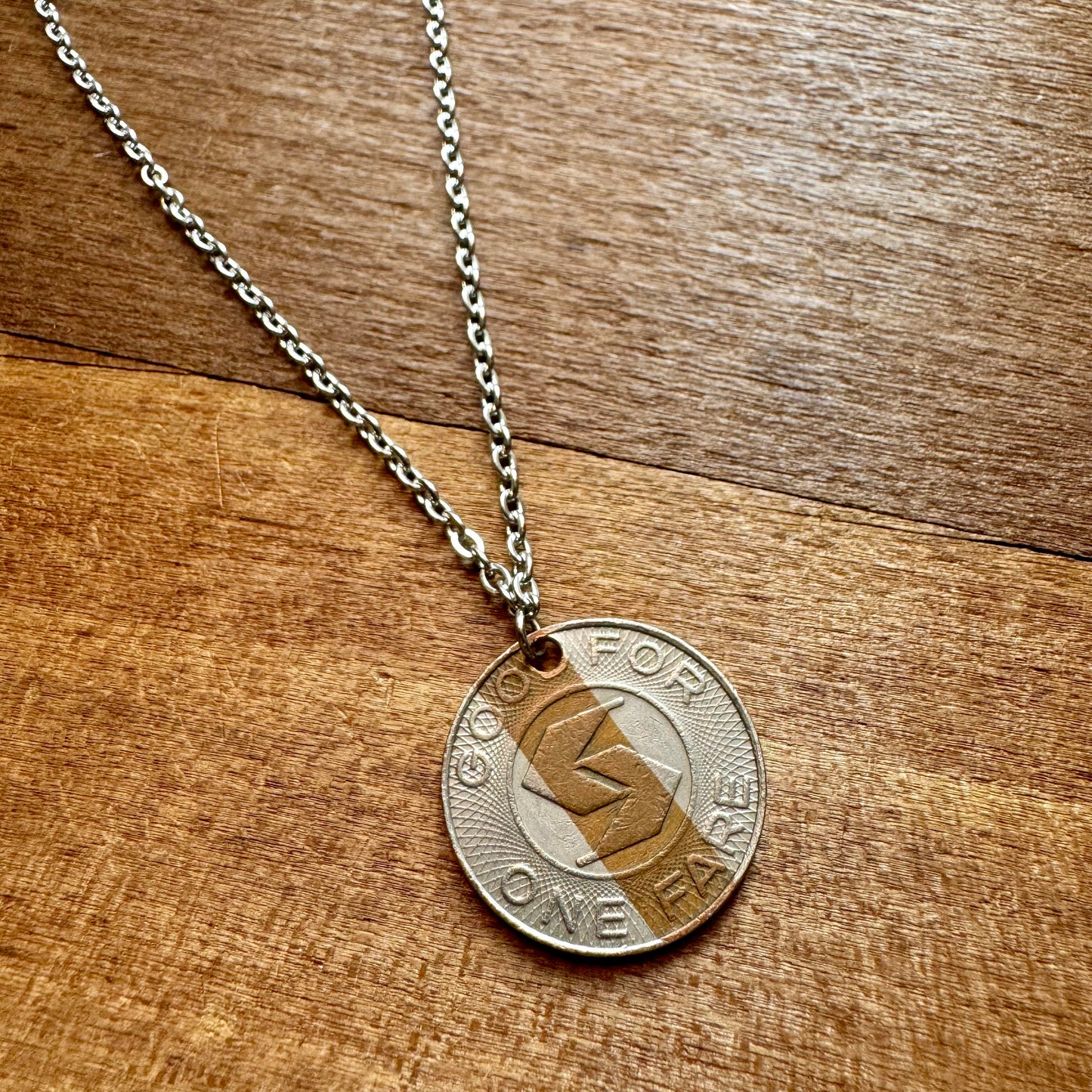 Introducing the SEPTA Token Necklace by INDICAN, featuring a sterling silver chain and a pendant crafted from an authentic metal SEPTA token inscribed with "GOOD FOR ONE FARE," elegantly displayed on a wooden background.