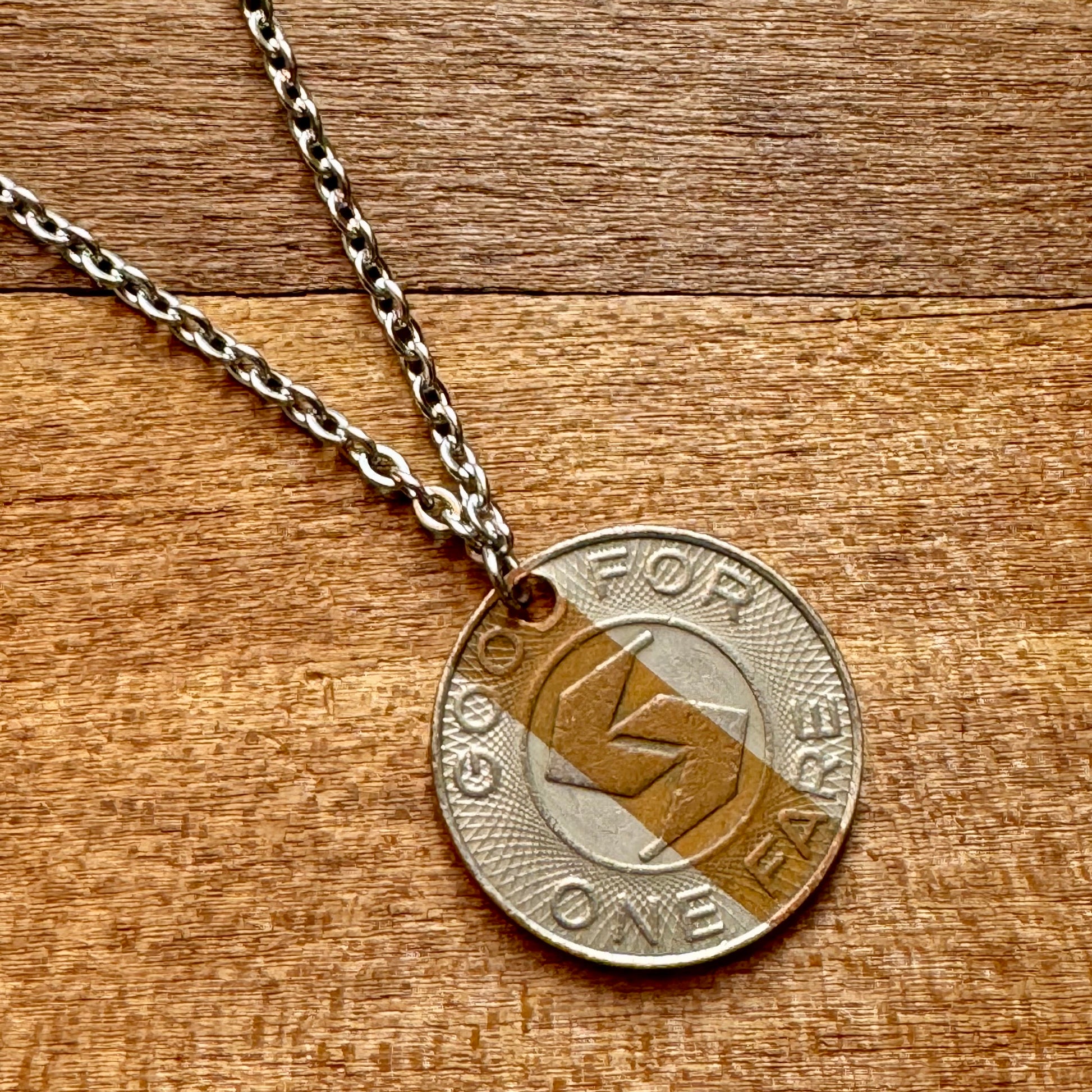 The INDICAN Septa Token Necklace features an old SEPTA token inscribed with "GOOD FOR ONE FARE" on a sterling silver chain, set against a wooden background.