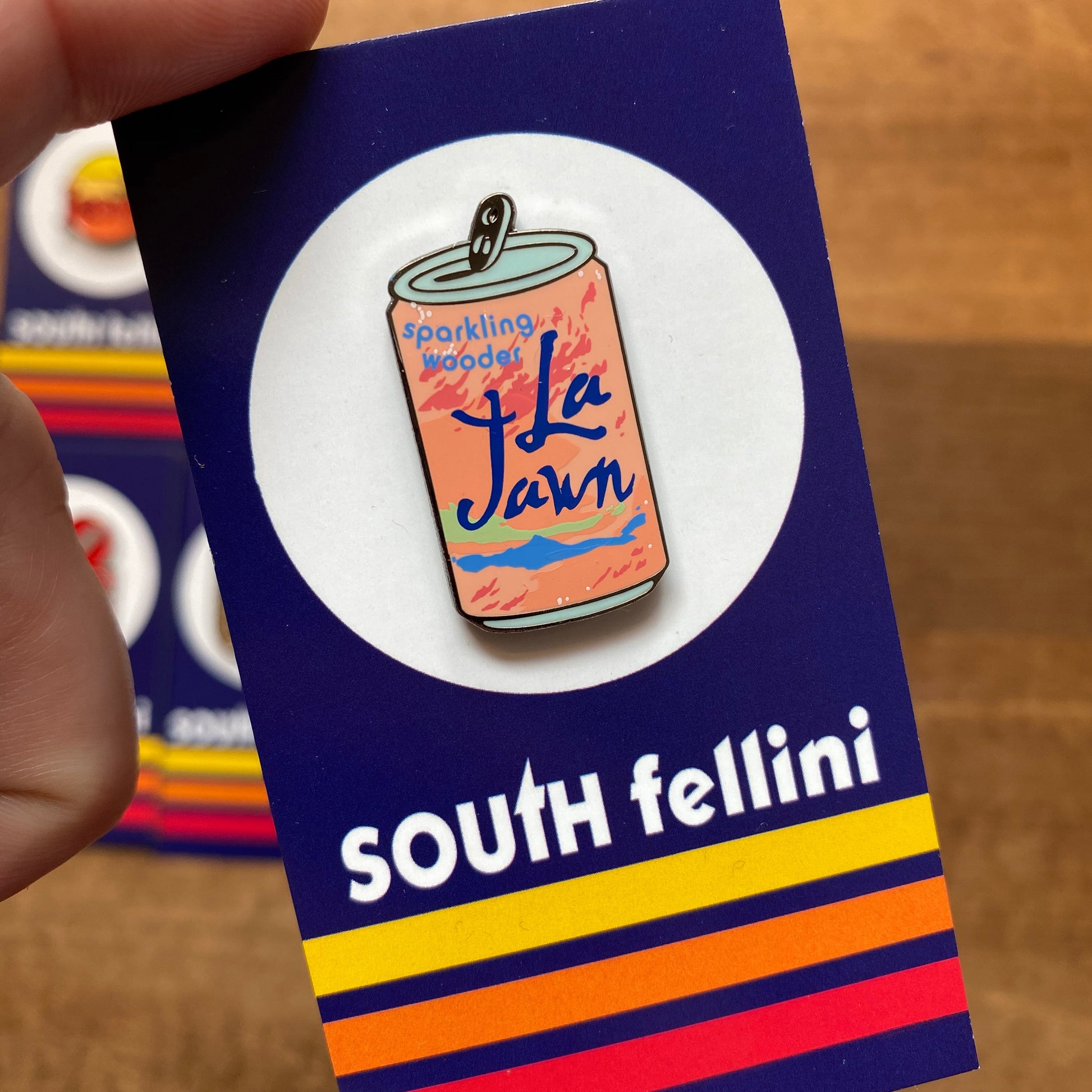 A hand holds one of the Philly-Themed Enamel Pins from South Fellini, designed to resemble a can labeled "Sparkling Wooter La Jawn," a charming nod to Philly icons. The pin is attached to a card featuring the text "South Fellini" and colorful stripes at the bottom.