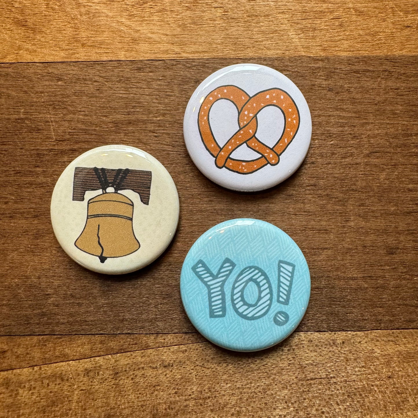 Three round pins with different designs: a coffee pot, a Philly Pretzel magnet, and the word "yo!" on a wooden surface from the exit343design Philly Magnet Set.