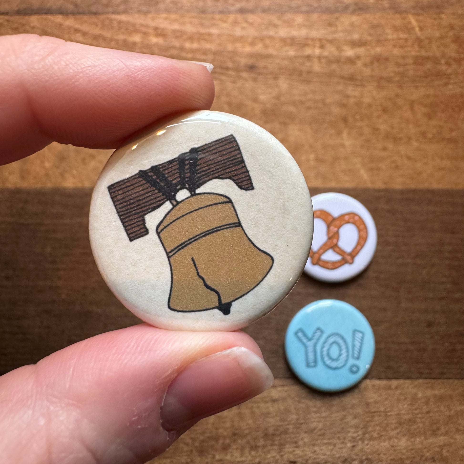 A hand holding a Philly Magnet Set by exit343design with an illustration of the liberty bell, next to two other smaller magnets with a Philly Pretzel and the word "yo!" on a wooden surface.