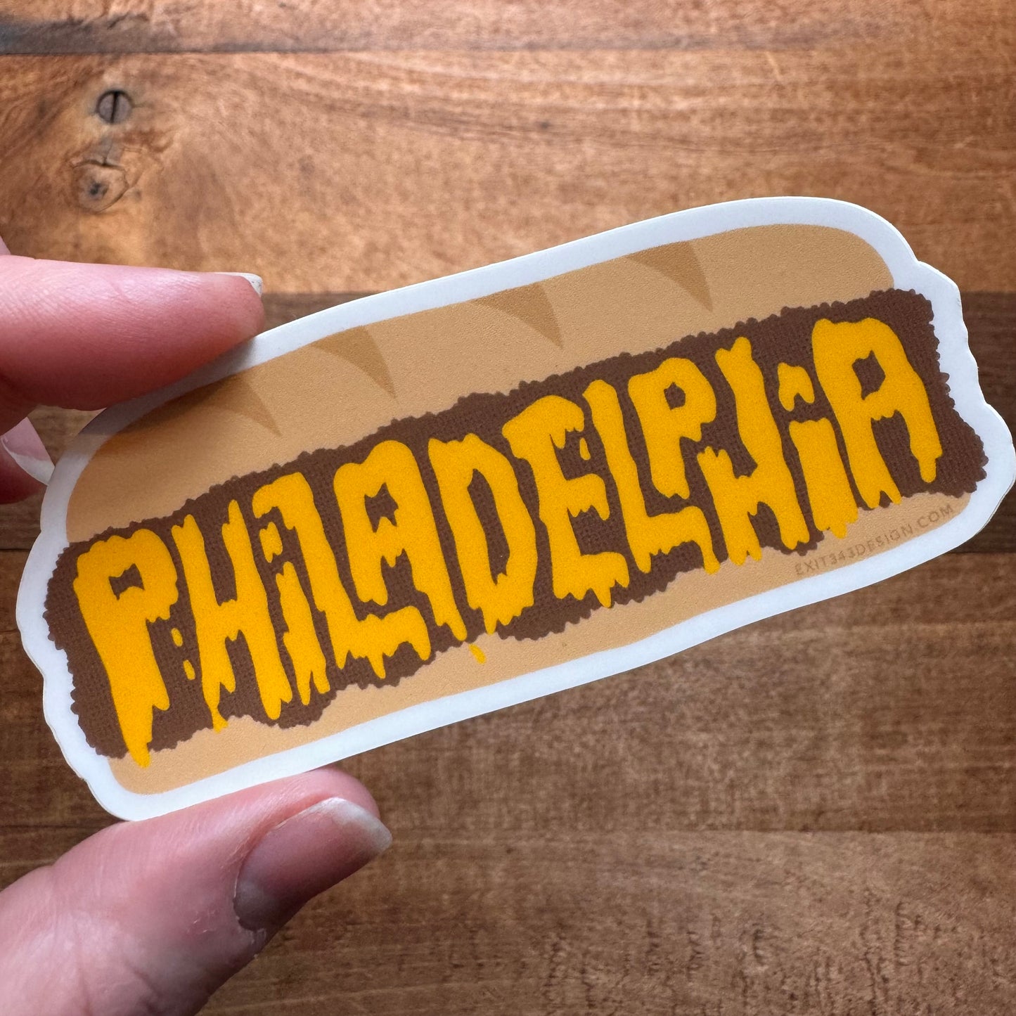 A hand holding a Philly Stickers with the word "Philadelphia" in a stylized font on a wooden surface, made by exit343design.