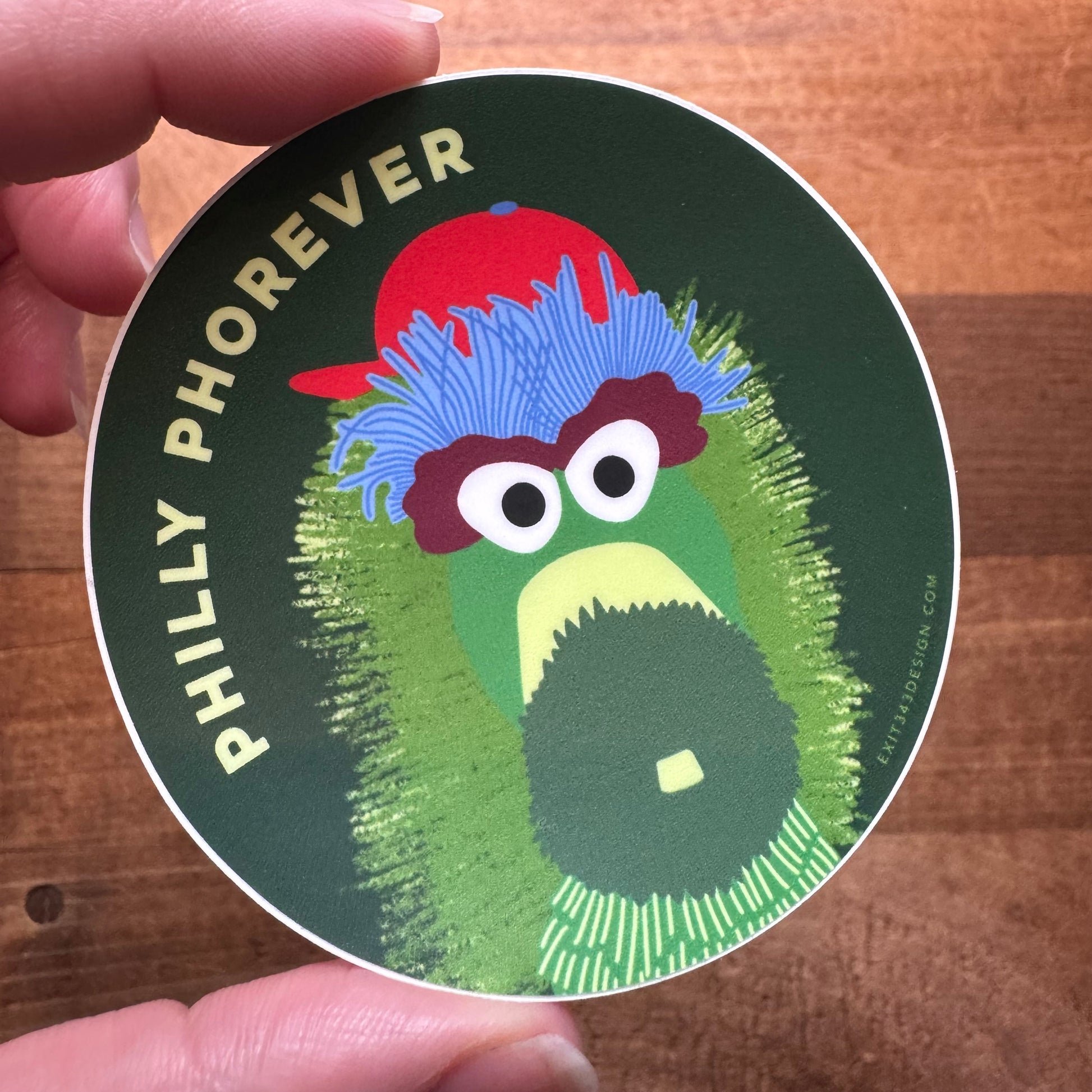 A hand holding a circular vinyl Philly Sticker with a stylized illustration of the Philadelphia Phillies mascot, the Phillie Phanatic, and the text "Philly Phorever" made by exit343design.