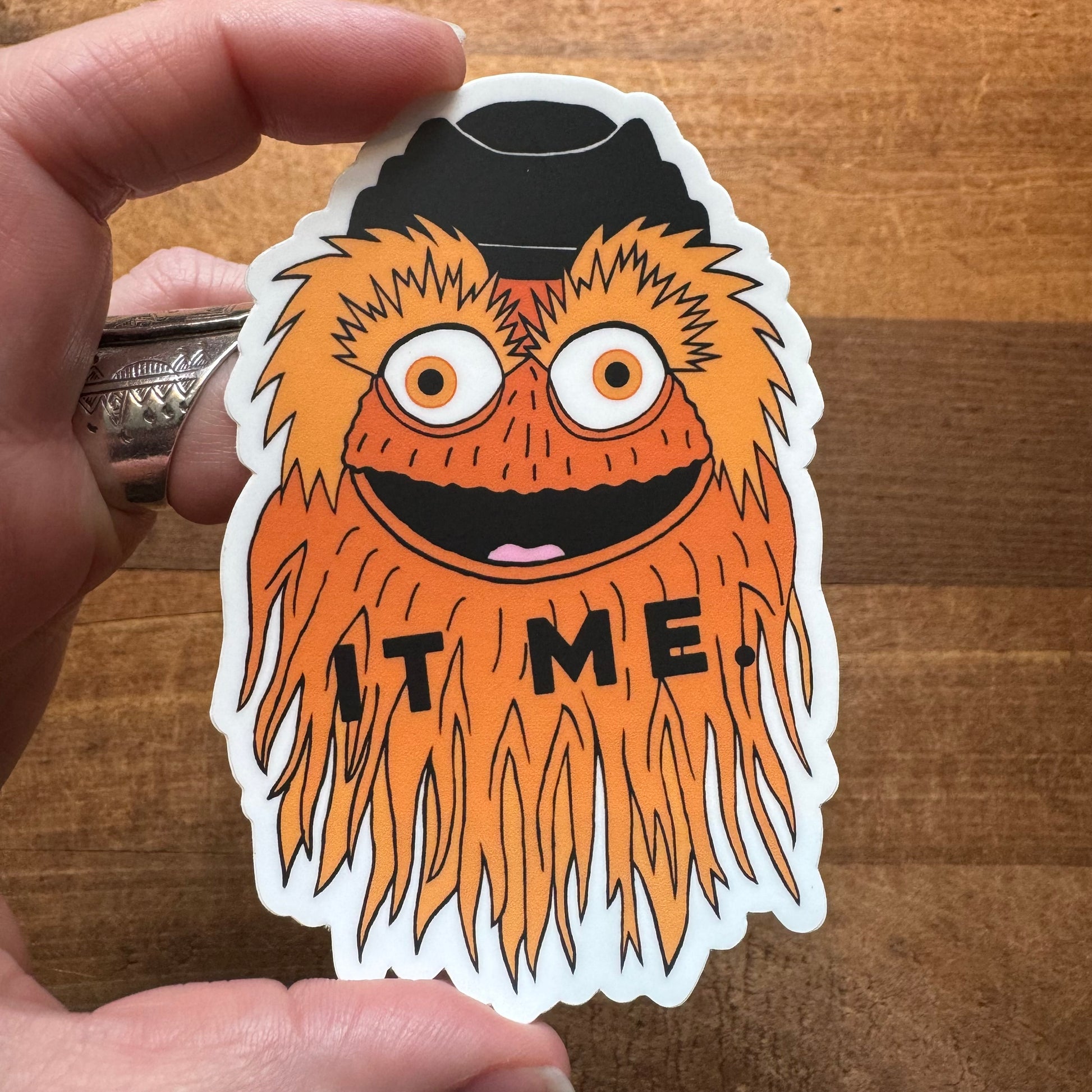 A hand holding a Philly-themed vinyl sticker of a whimsical orange character with the word "time" on its chest, made by exit343design.