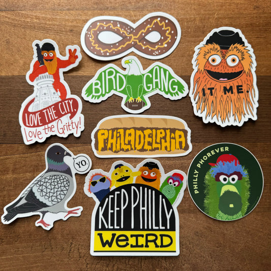 Assortment of colorful, exit343design Philly Stickers including mascots such as Gritty and text related to cheesesteaks, displayed on a wooden surface.