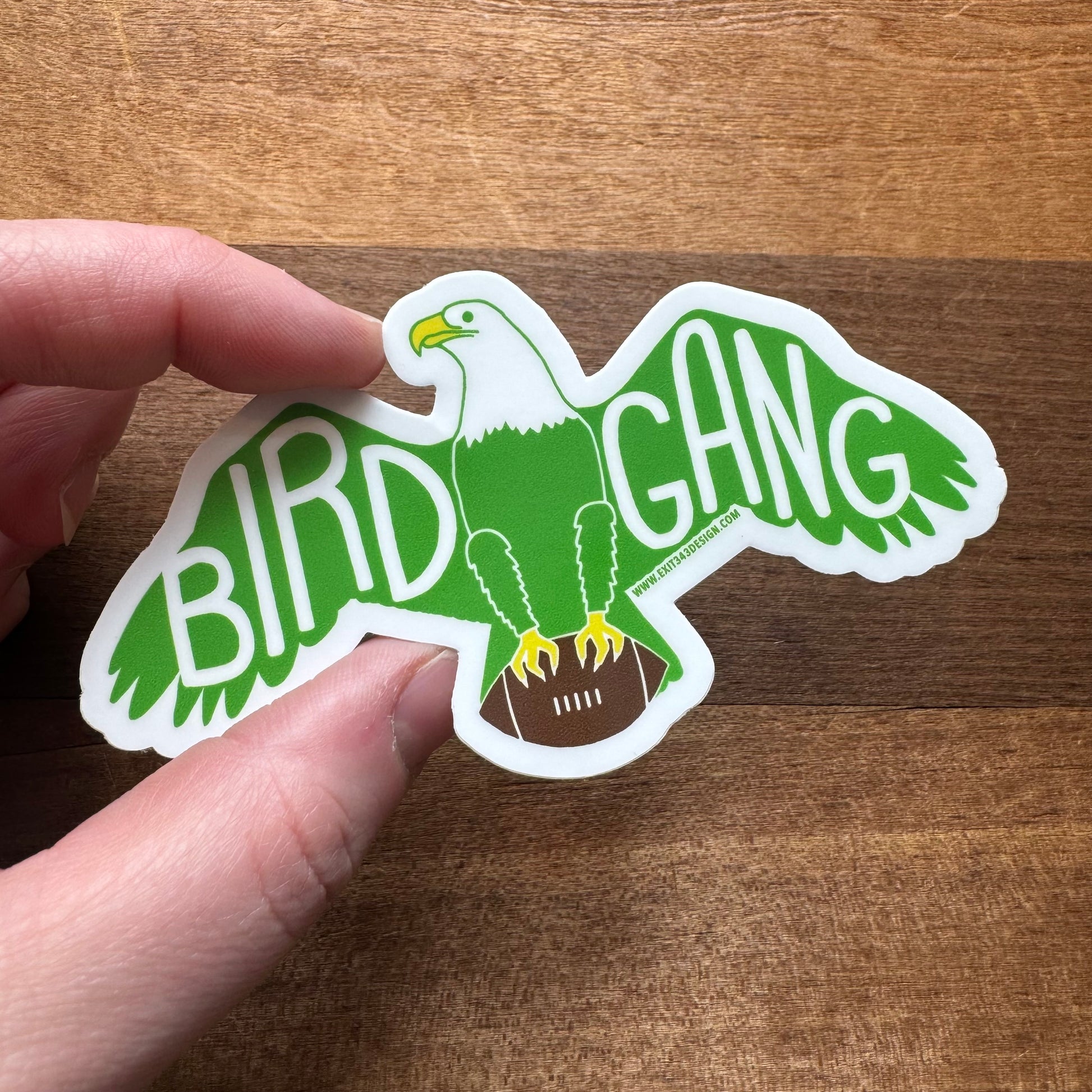 A hand holding an exit343design Philly Stickers shaped like an eagle with the words "bird gang" written in green on a wooden background.