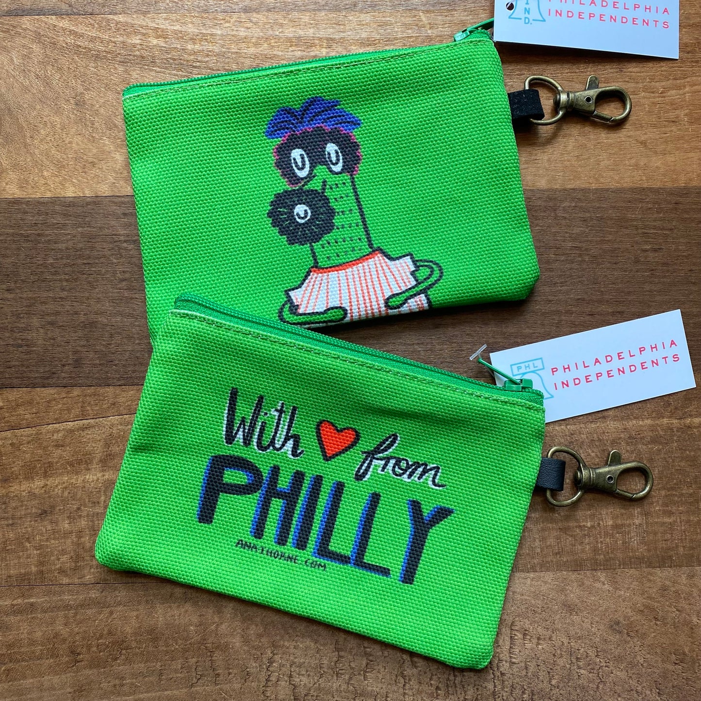 Two green Gritty & Phanatic zipper pouches with embroidery, one featuring a character and the other the text "with love from philly," on a wooden background. By Ana Thorne.