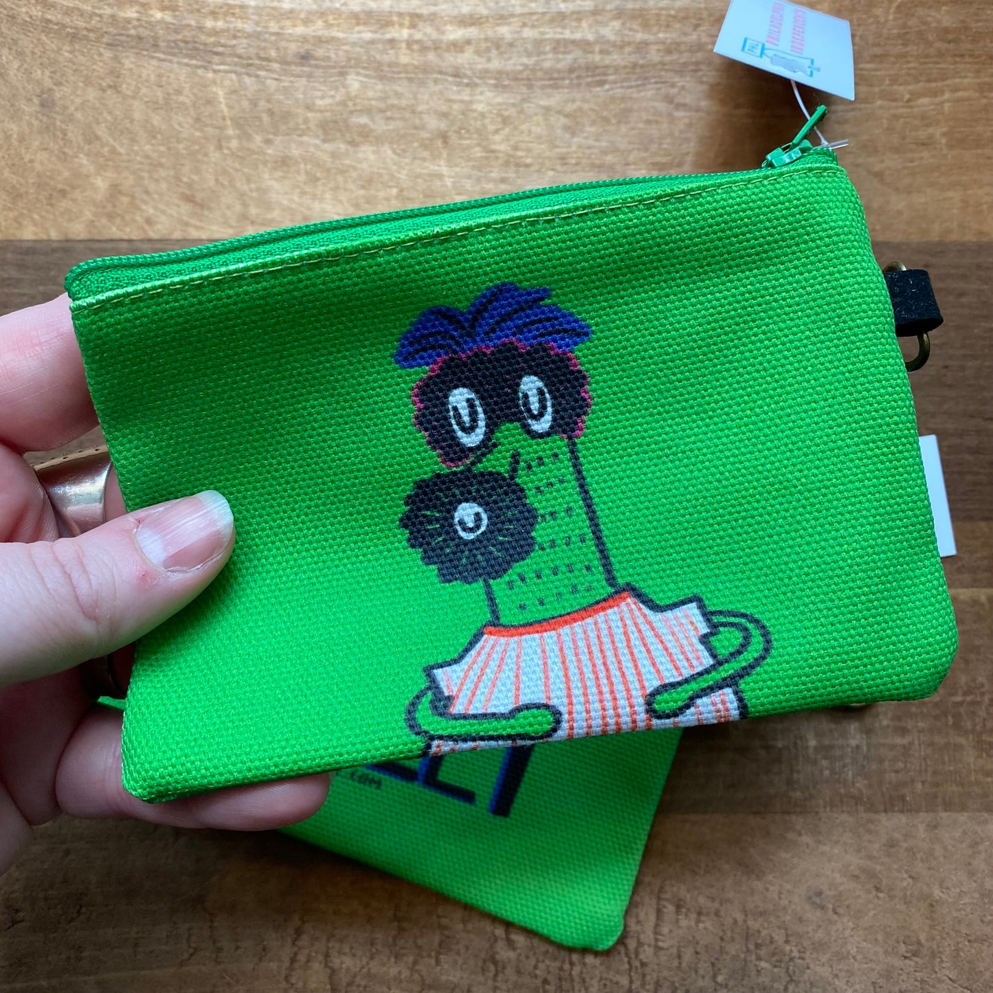 A hand holding a green Gritty & Phanatic zipper pouch with a cartoon character design on a wooden background by Ana Thorne.