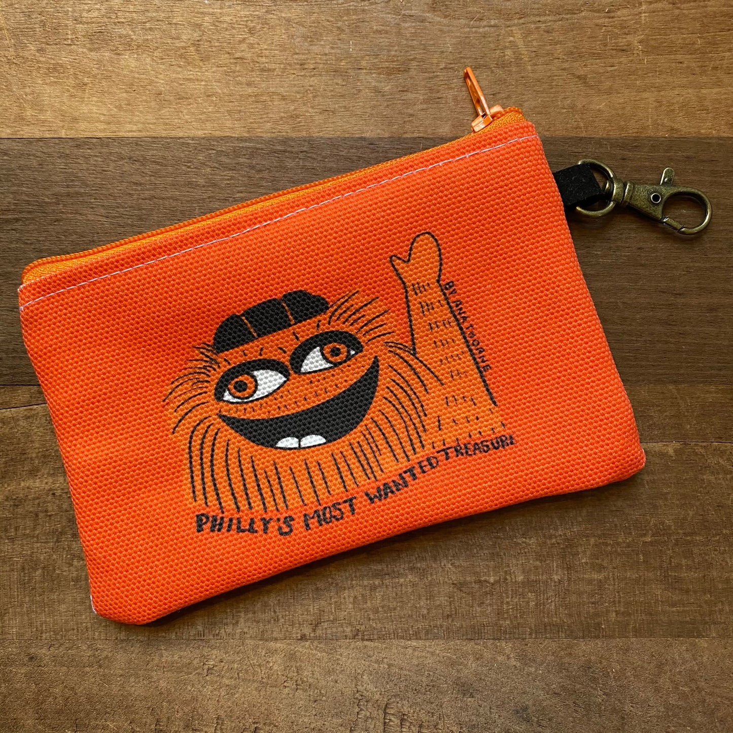 Orange Gritty & Phanatic Zipper Pouch with an illustration of a whimsical cartoon cat face and text that reads "Philly’s Most Wanted Treasure" by Ana Thorne.