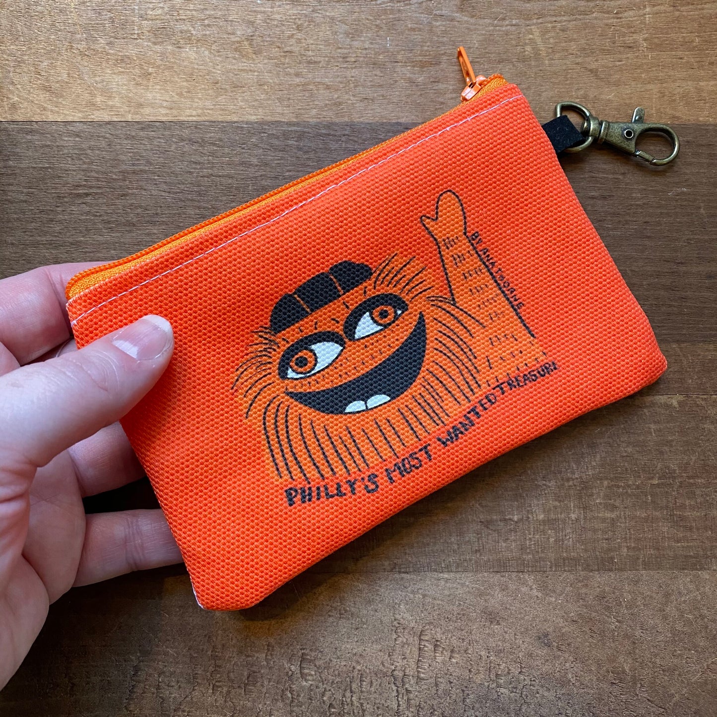 A hand holding a Gritty & Phanatic Zipper Pouch with a cartoonish figure and text "Philly’s Most Wanted Treasure" by Ana Thorne.
