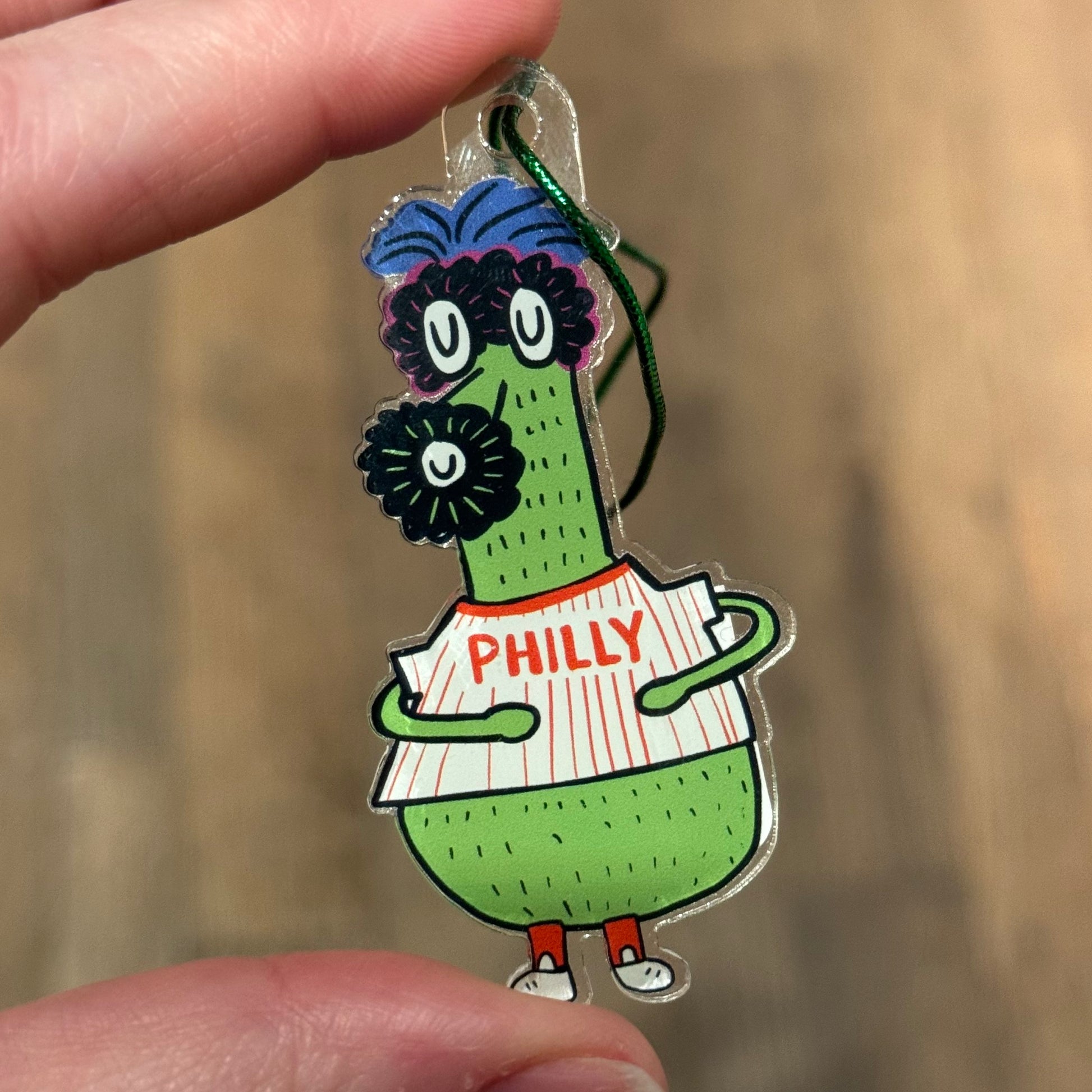 A hand holds a Philly Acrylic Ornament featuring a green cartoon character wearing a jersey labeled "PHILLY." The ornament, designed by Ana Thorne, has exaggerated facial features and a blue and purple pom-pom hat, embodying true Philly pride.