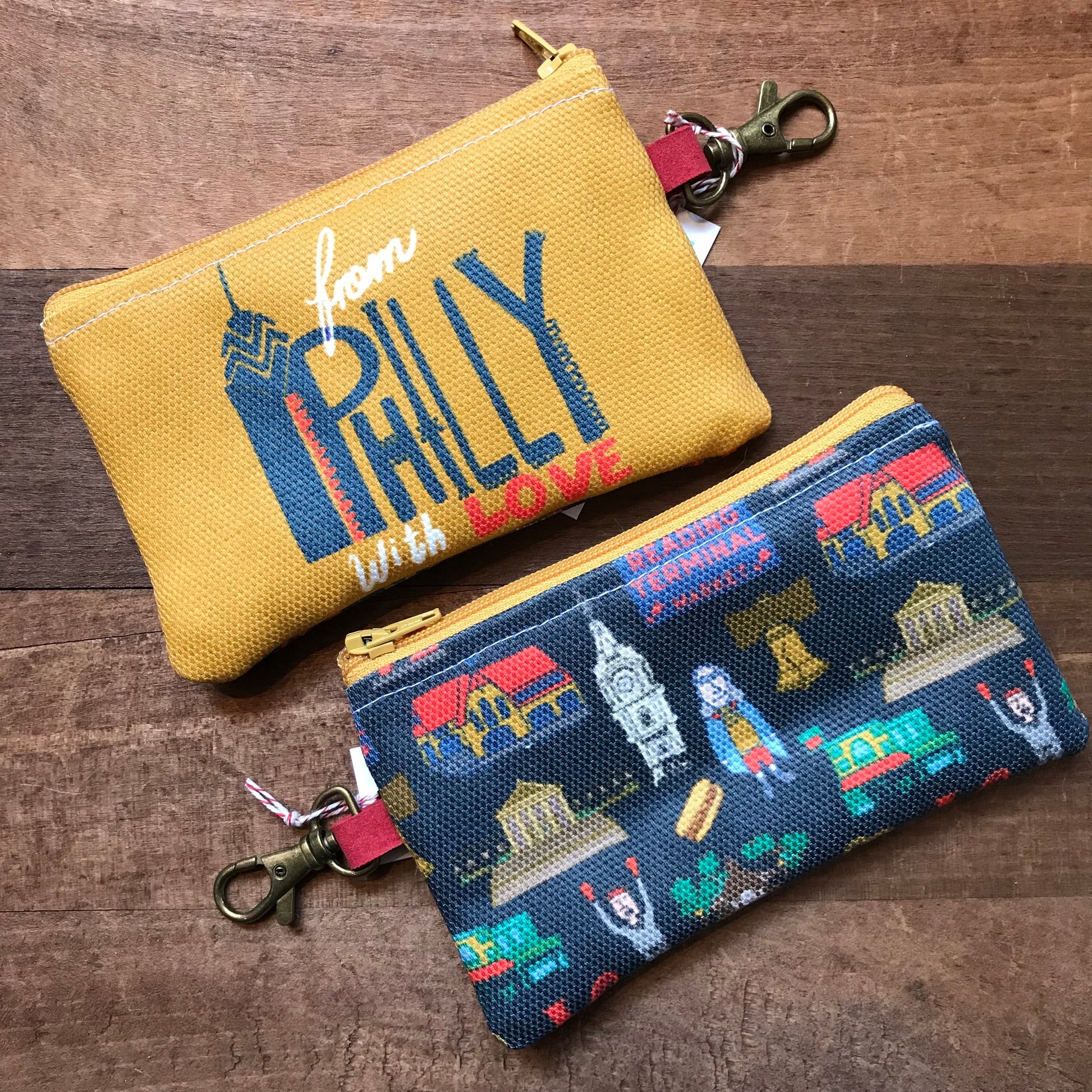 Two Philly Zipper Pouches with Philadelphia-themed designs on a wooden background by Ana Thorne.