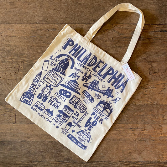 A lightweight canvas Philadelphia Icons Tote Bag featuring illustrations and text related to Philadelphia landmarks and culture, designed by Ana Thorne, on a wooden surface.