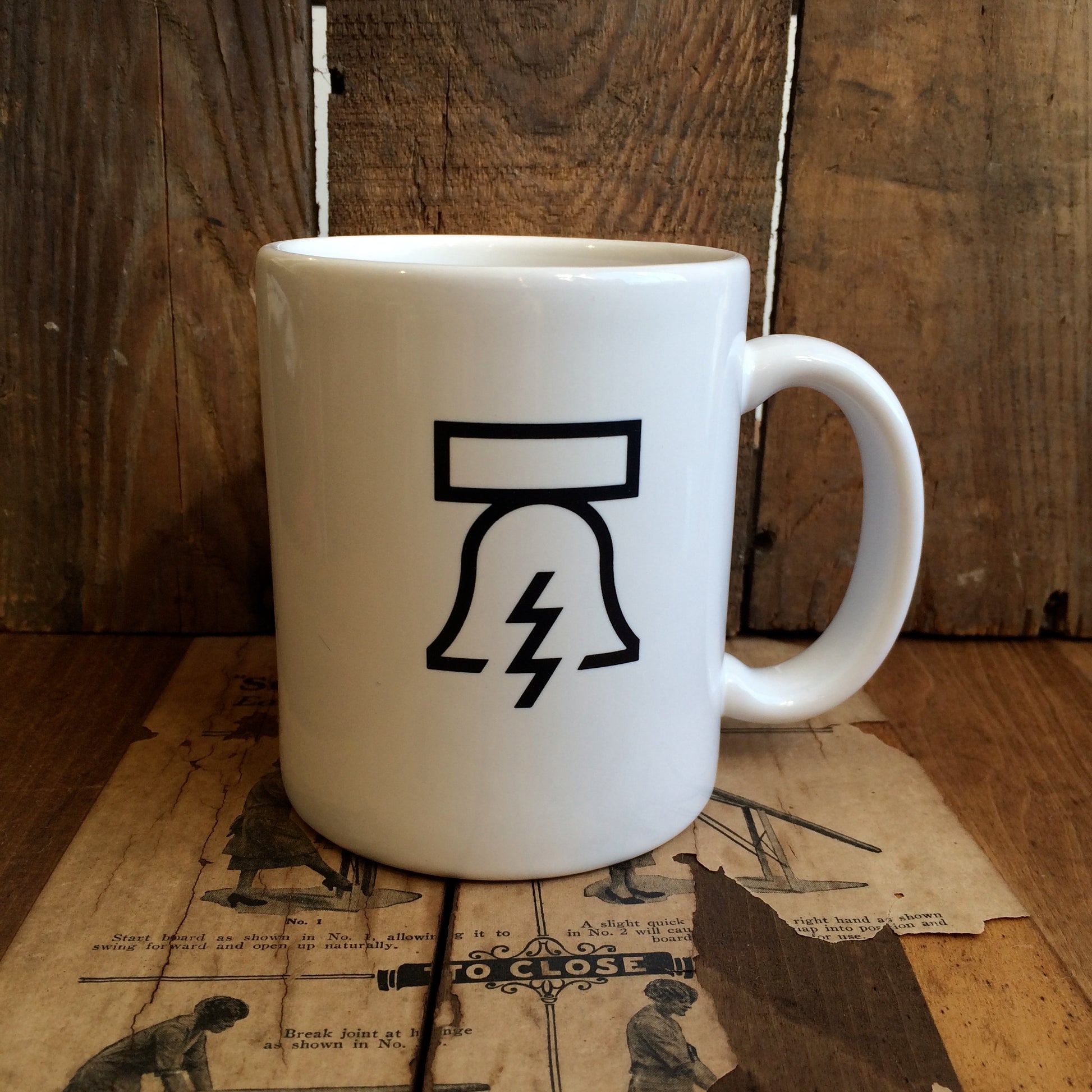 A white Bell & Bolt Mug by Philadelphia Independents featuring a simplistic black design of the iconic bell with a lightning bolt is placed on a wooden surface.