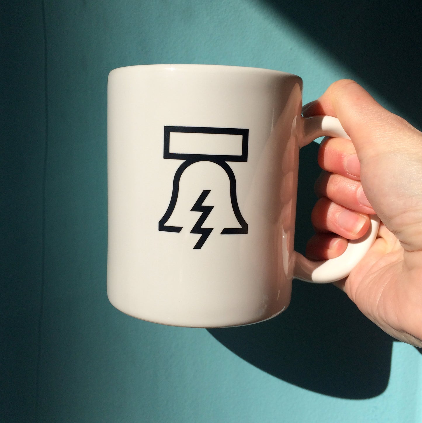 A hand holding a white Bell & Bolt Mug by Philadelphia Independents with a black graphic design of a bell featuring a lightning bolt on it.