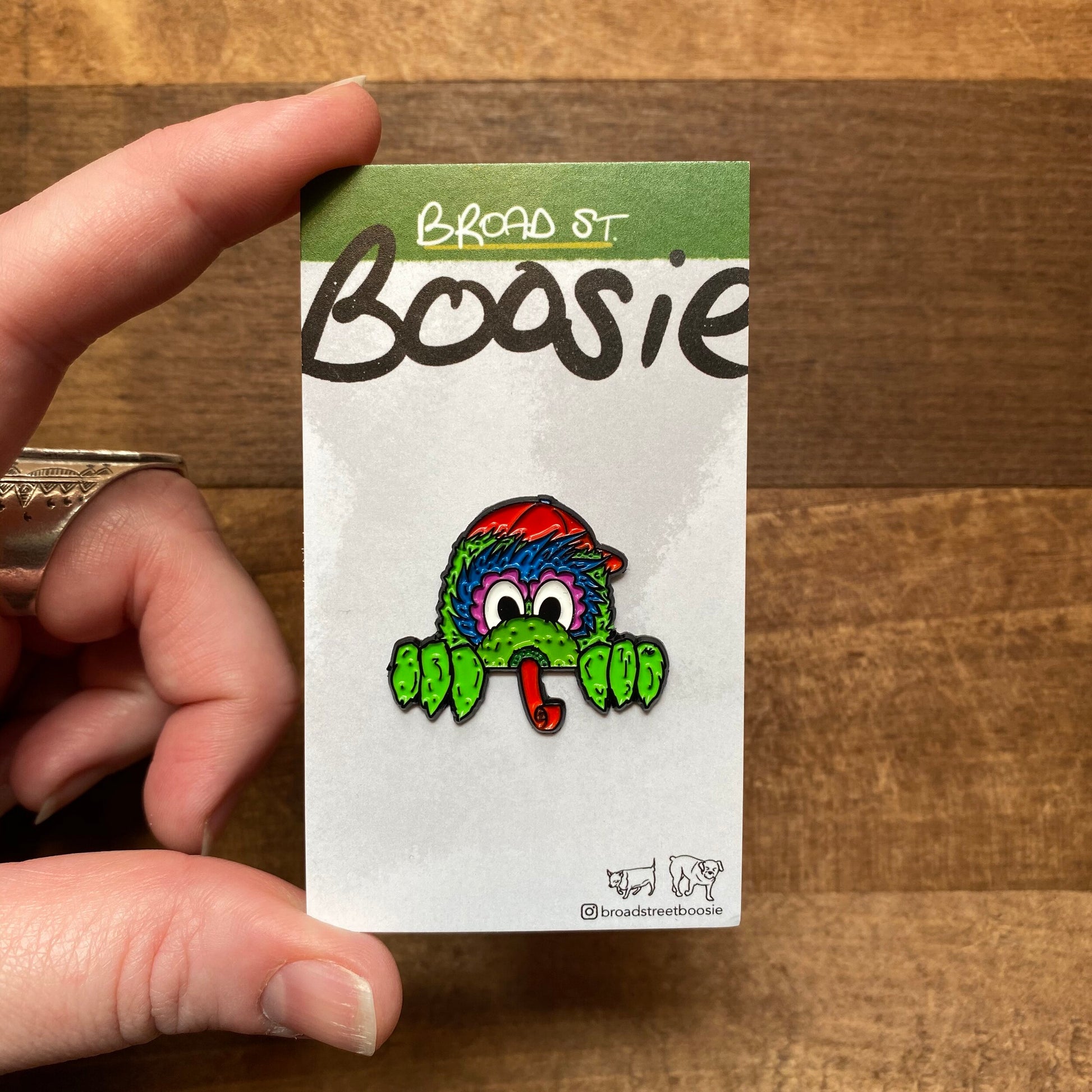 A hand holding a Gritty & Phanatic Enamel Pin designed with a colorful, cartoonish character against a notepad with "Broad Street Boosie" handwritten at the top.