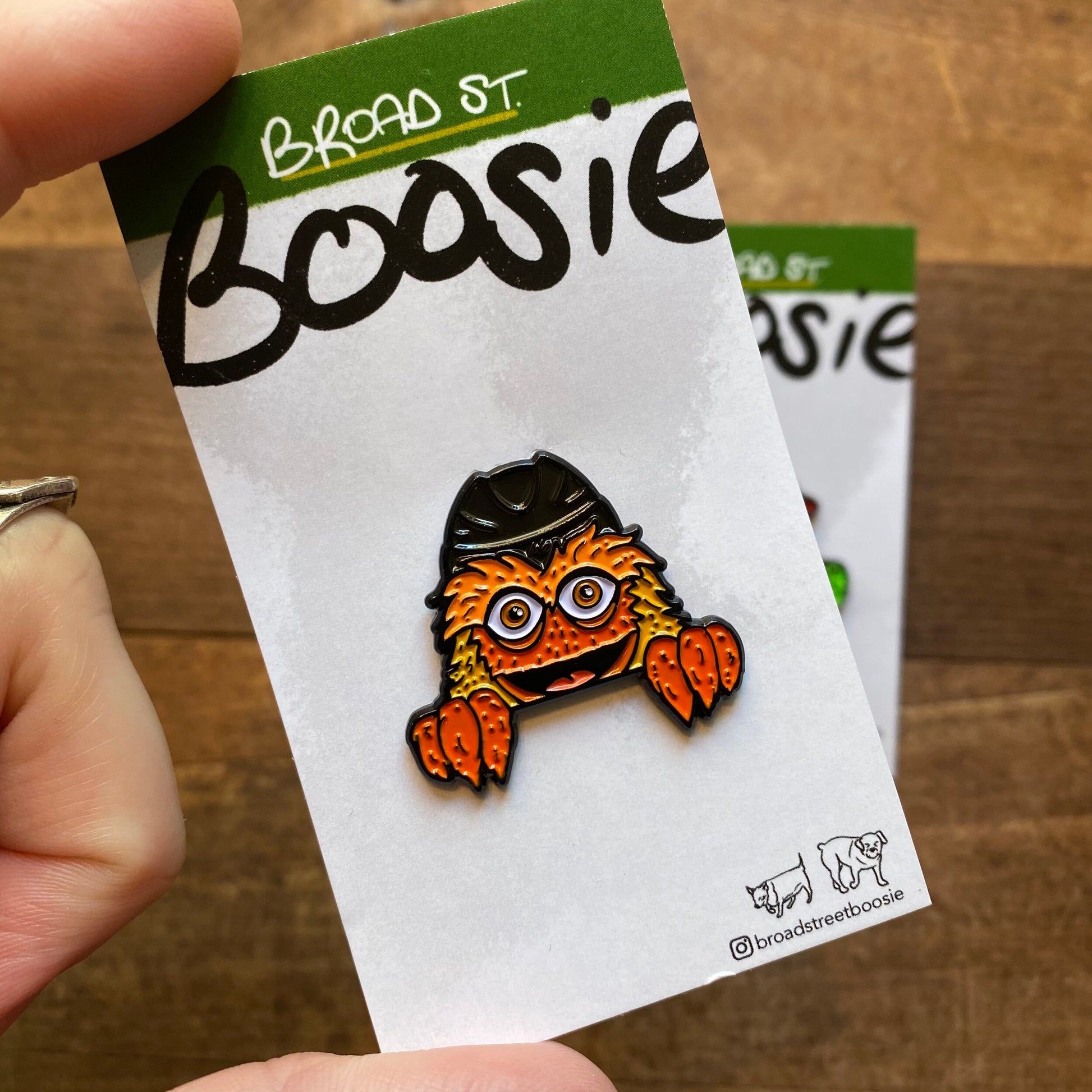 A hand holding a Gritty & Phanatic enamel pin featuring an orange, hairy monster character with large eyes displayed on a card labeled "Broad Street Boosie".