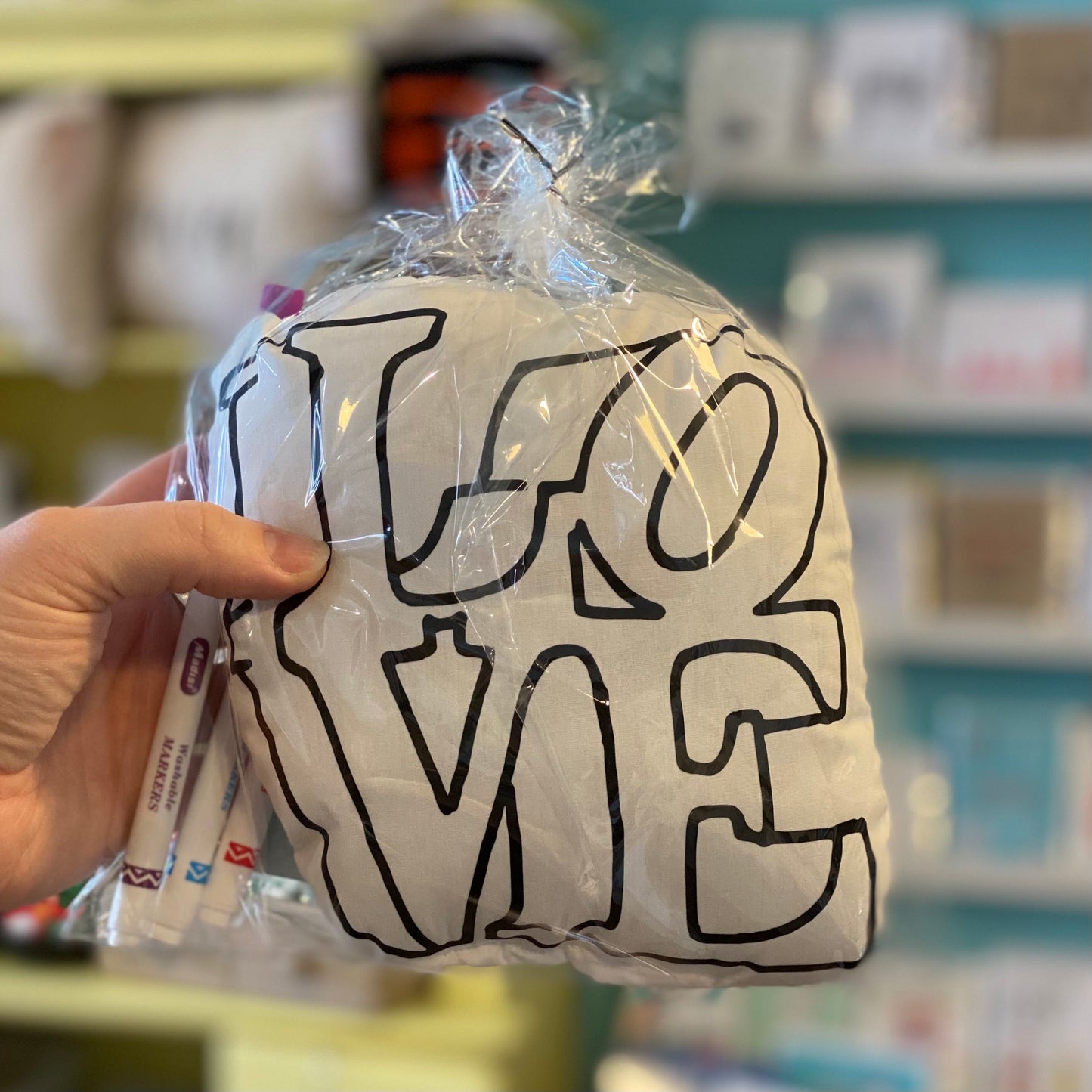 A hand holding a wrapped Doodle Jawnz Washable Kits with the word "Love Statue" in bold, stylized letters, in front of a blurred background of colorful shelves.