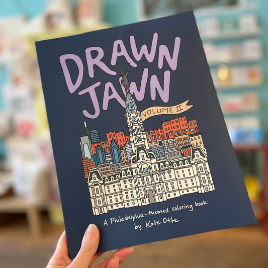 A hand holding the "Drawn Jawn Coloring Book," an original drawings Philadelphia-themed coloring book by Kate Otte, against a blurred background.