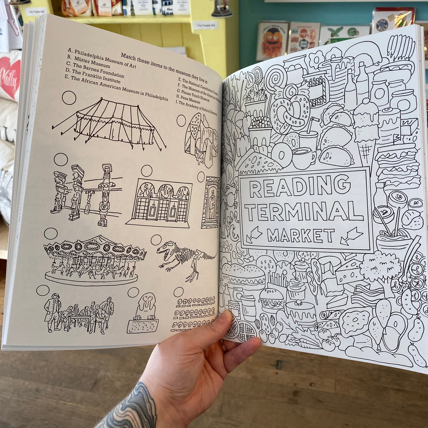 A person holding an open Drawn Jawn Coloring Book by Kate Otte featuring a page with original drawings related to Reading Terminal Market.
