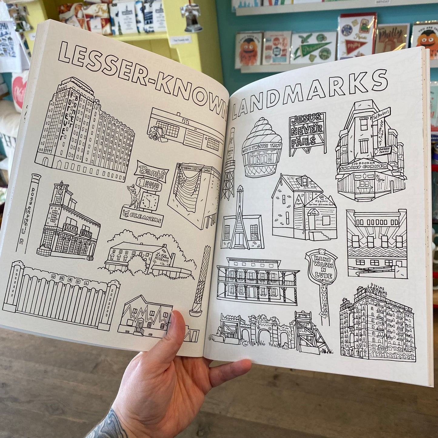 A person holding open a Drawn Jawn Coloring Book titled "lesser-known landmarks" featuring original drawings of various architectural structures by Kate Otte.