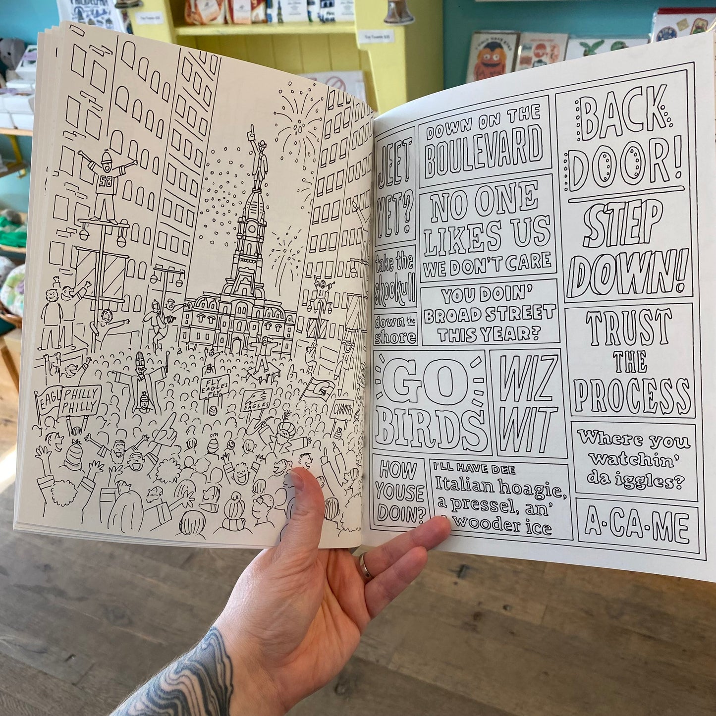A person holding open a Drawn Jawn Coloring Book with detailed black and white original drawings of Philly places and playful text by Kate Otte.