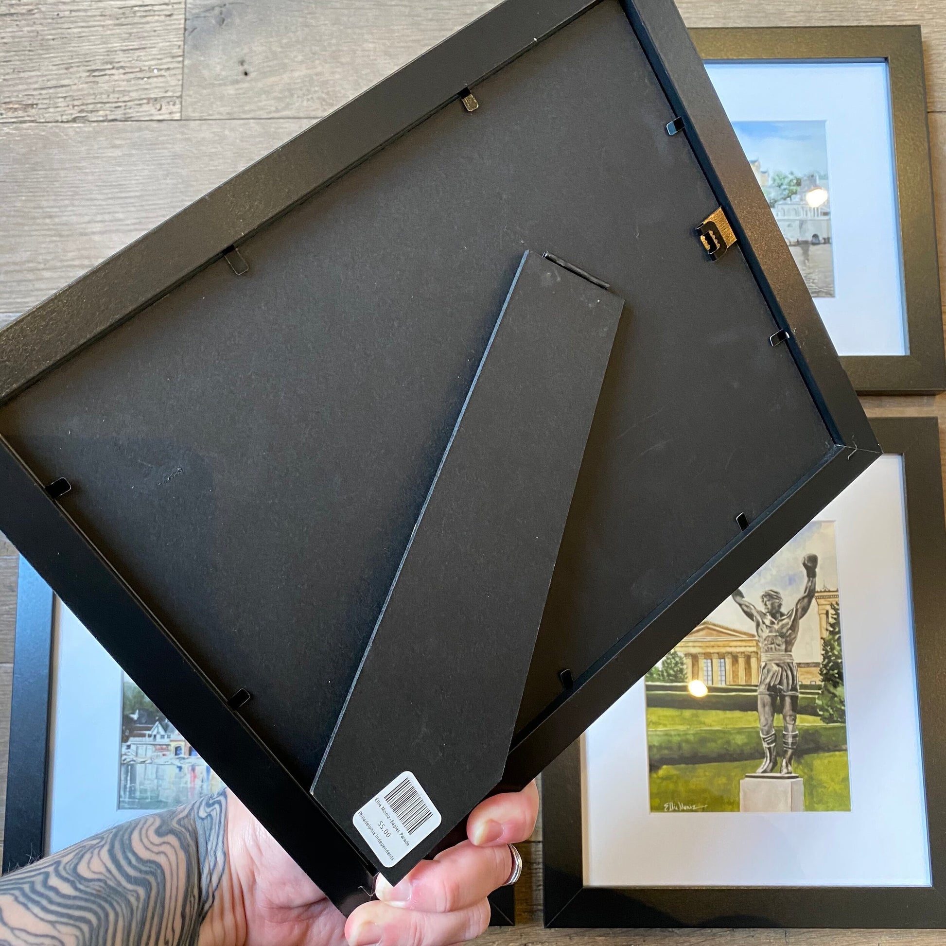 A person holding the back of a black picture frame with other framed Ellie Moniz Philly Watercolor prints visible in the background.