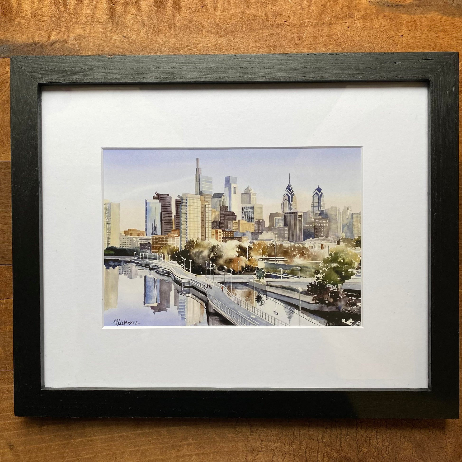 A framed Philly Watercolor Print of the Philadelphia skyline with prominent buildings and reflective water in the foreground, displayed on a wooden surface by Ellie Moniz.