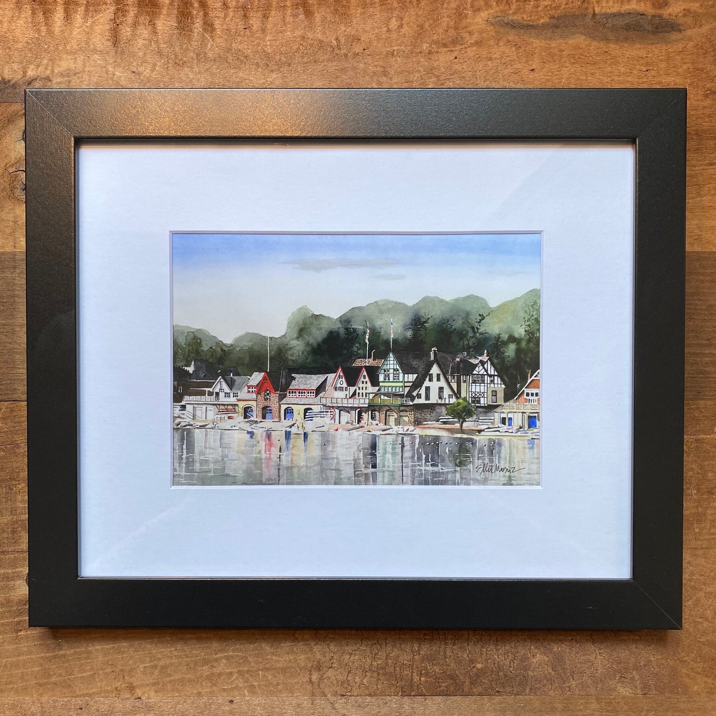 Framed Philly Watercolor print of a serene lakeside village with quaint houses and mountains in the background, displayed on a wooden surface by Ellie Moniz.