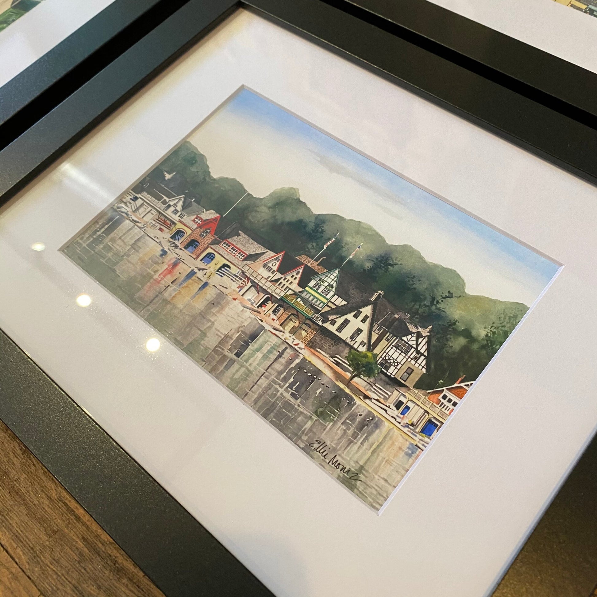 Ellie Moniz's Framed Philly Watercolor Prints of a quaint Philadelphia town with colorful houses and a mountain backdrop, displayed on a wooden table.