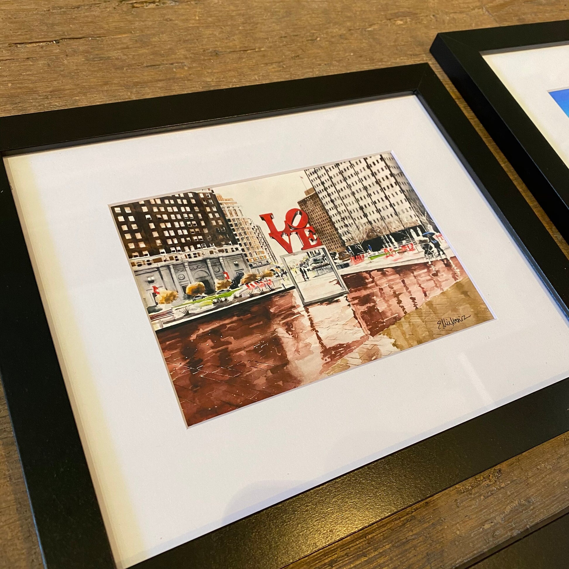 Framed Philly Watercolor Prints of a cityscape with buildings and a prominent red "love" sculpture, displayed on a wooden table by Ellie Moniz.