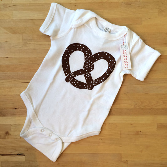 White cotton Pretzel Baby Onesie with a brown Philly pretzel heart design, displayed on a wooden floor by exit343design.