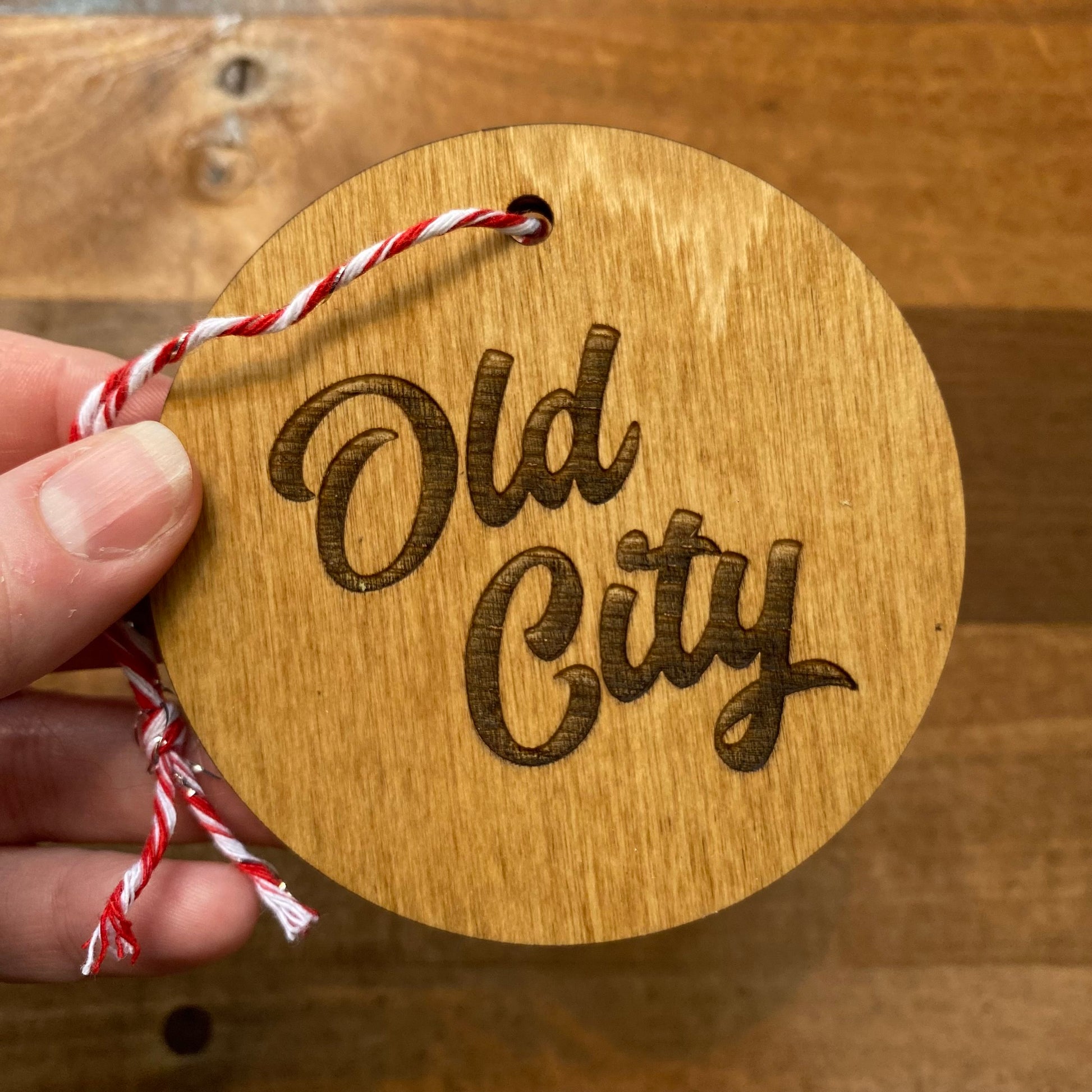 A hand holding a laser-etched Philly Wood Ornament disc with "Old City" engraved on it, attached to a red and white string. (Brand: Frankadelphia)