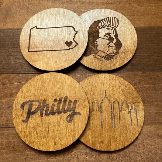 Four Mixed Philly Round Wood Coasters with engravings related to Philadelphia, including the state of Pennsylvania with a heart, a depiction of Ben Franklin, the word "Philly," and the city's skyline by Frankadelphia.