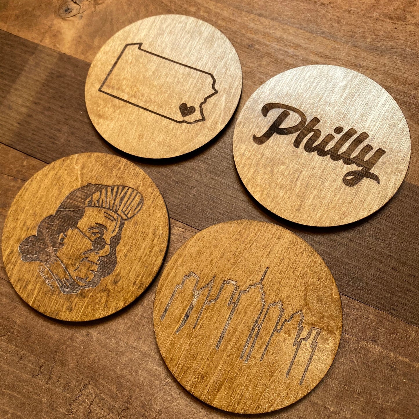 Frankadelphia's Mixed Philly Round Wood Coasters with engravings of notable symbols: the state of Pennsylvania with a heart, the word "Philly," Ben Franklin's profile, and the Philadelphia skyline.