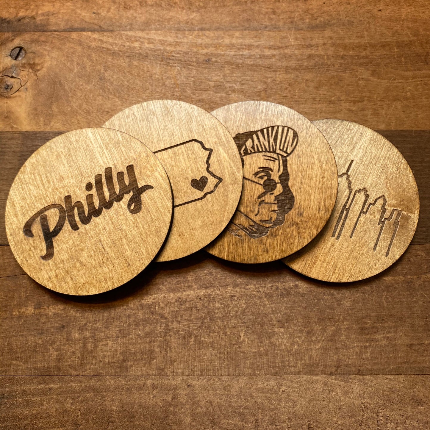 Four Frankadelphia Mixed Philly Round Wood Coasters with different engravings representing Philadelphia, including one with the city's nickname "Philly," a silhouette of the state of Pennsylvania with a heart, a depiction of Ben