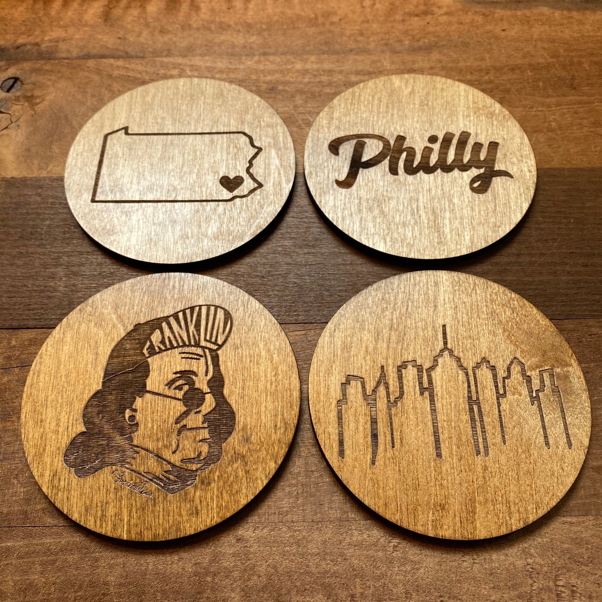 Four Frankadelphia Mixed Philly Round Wood Coasters with different engravings including the state outline with a heart, the word "Philly", Ben Franklin's face, and the city skyline.