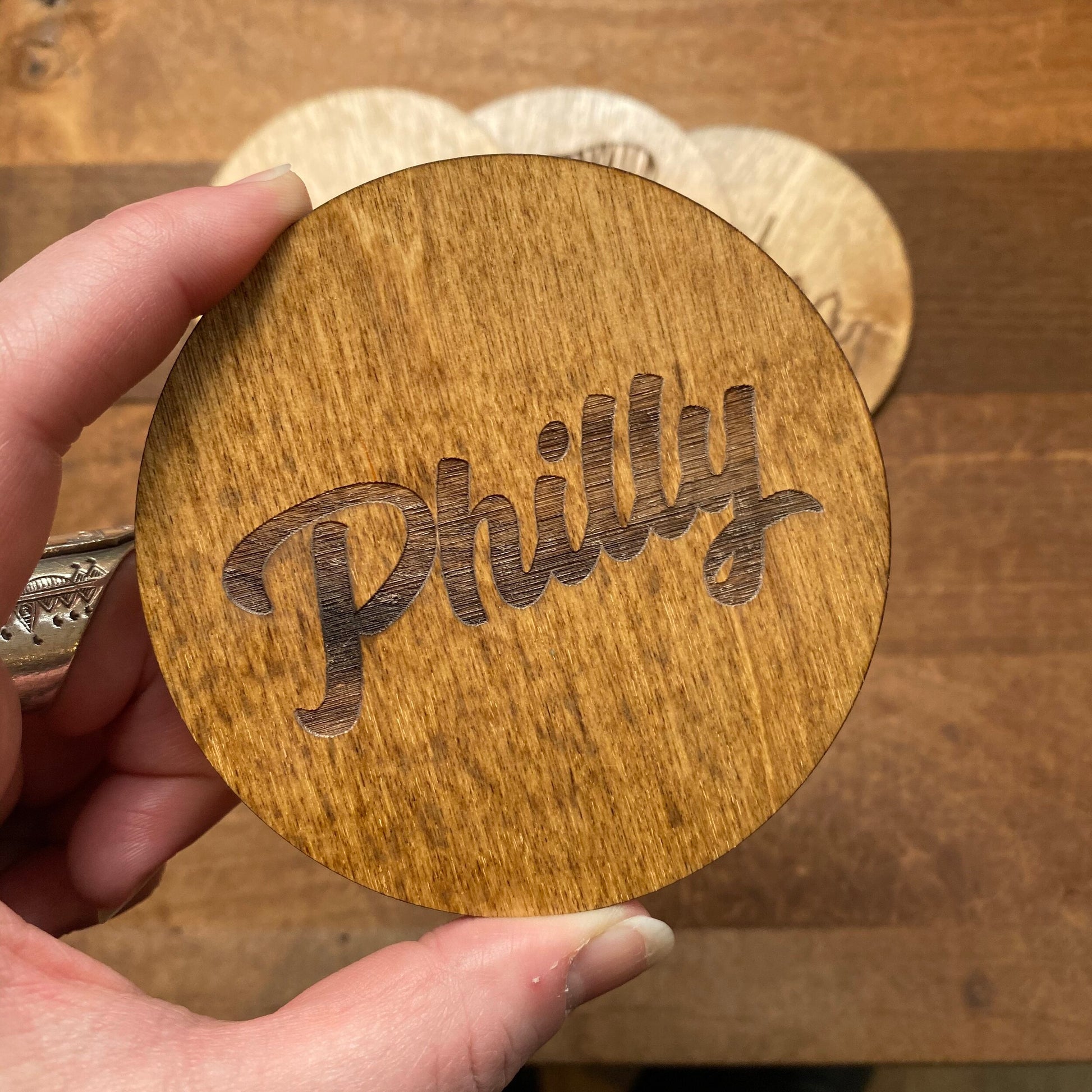 A hand holding a Mixed Philly Round Wood Coaster from Frankadelphia with the word "philly" engraved on it.