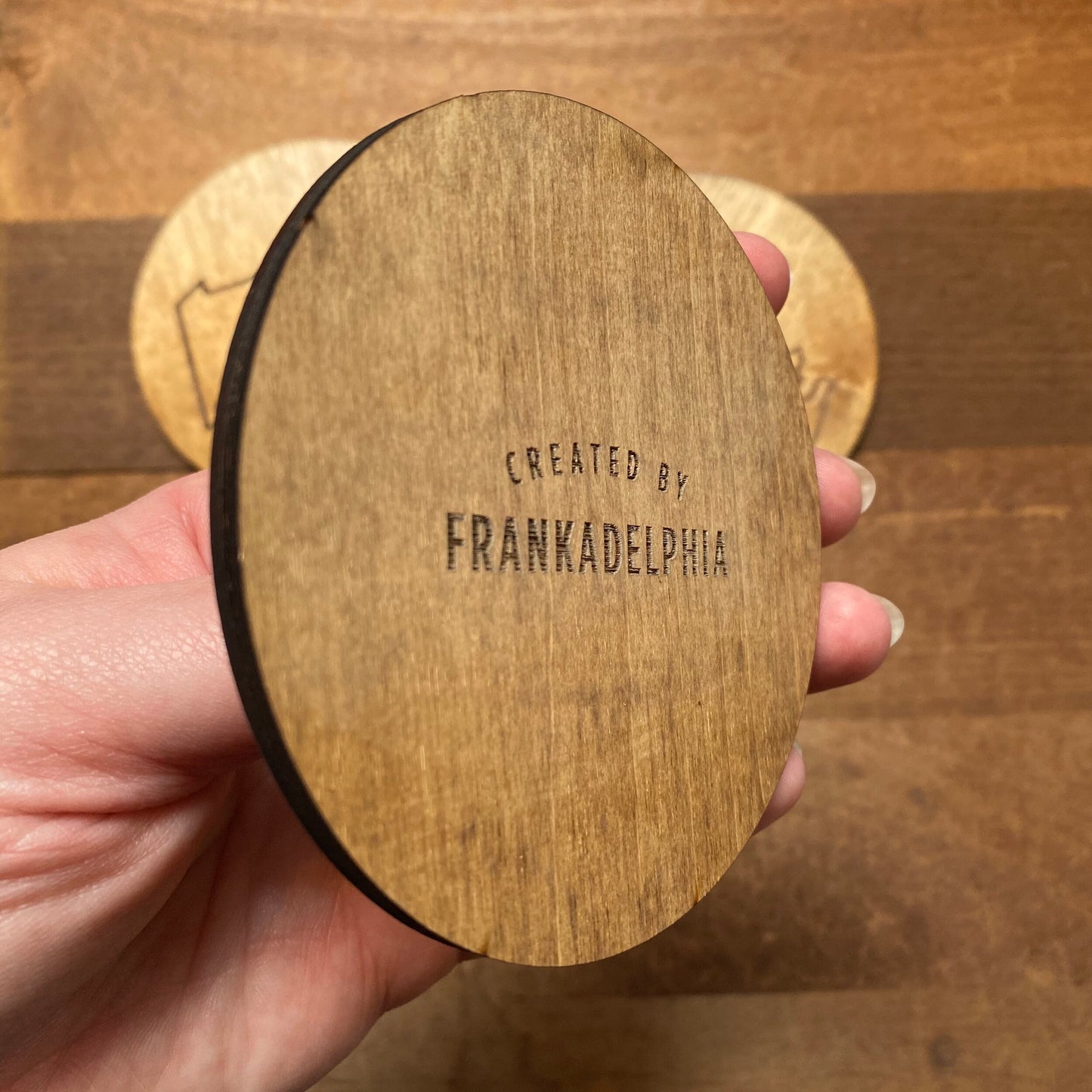 A hand holding a pack of Mixed Philly Round Wood Coasters with the words "created by Frankadelphia" burnt into them.